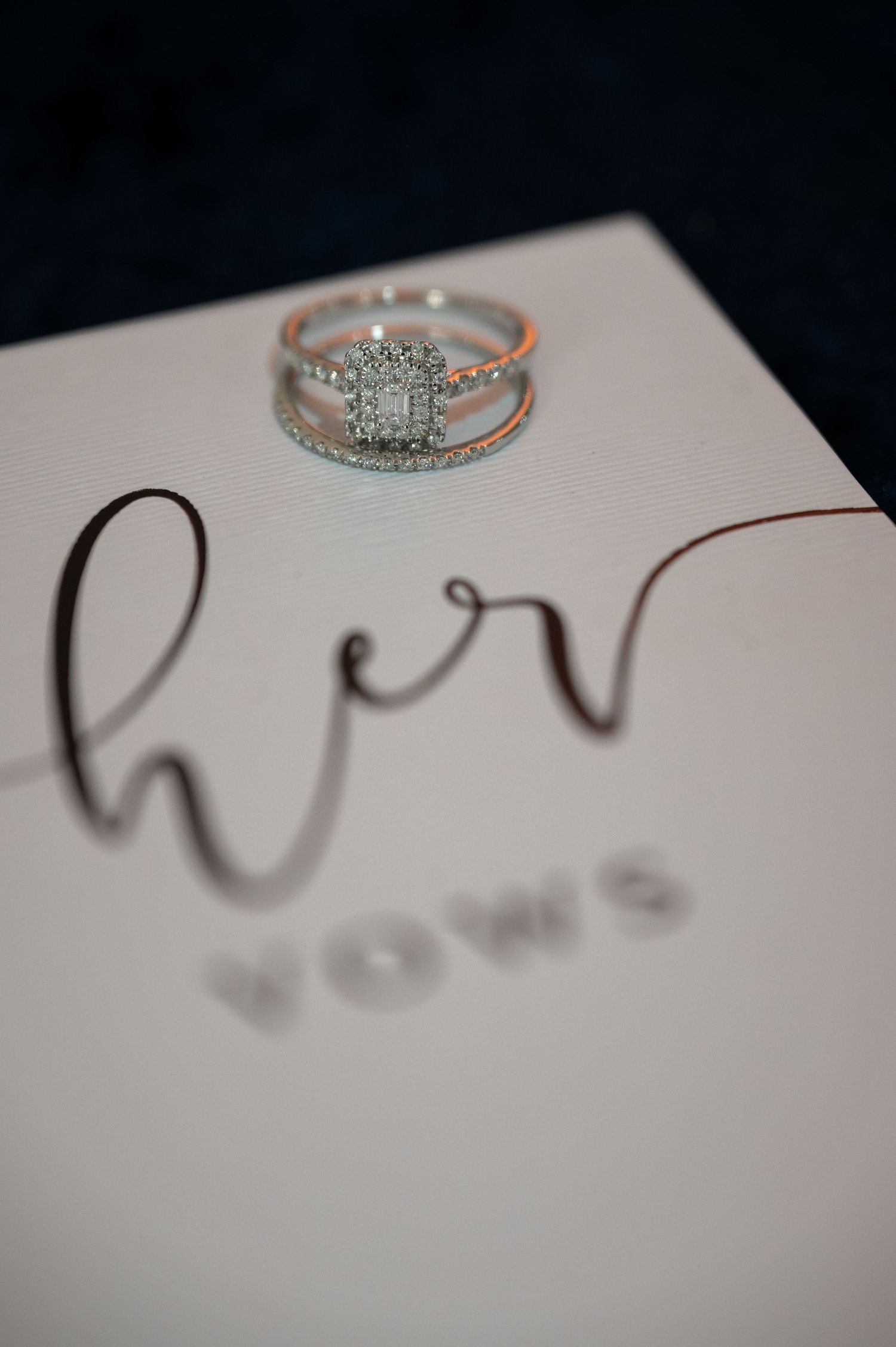 Diamond engagement ring set atop a white her vows book, with elegant script lettering and dark blue background.