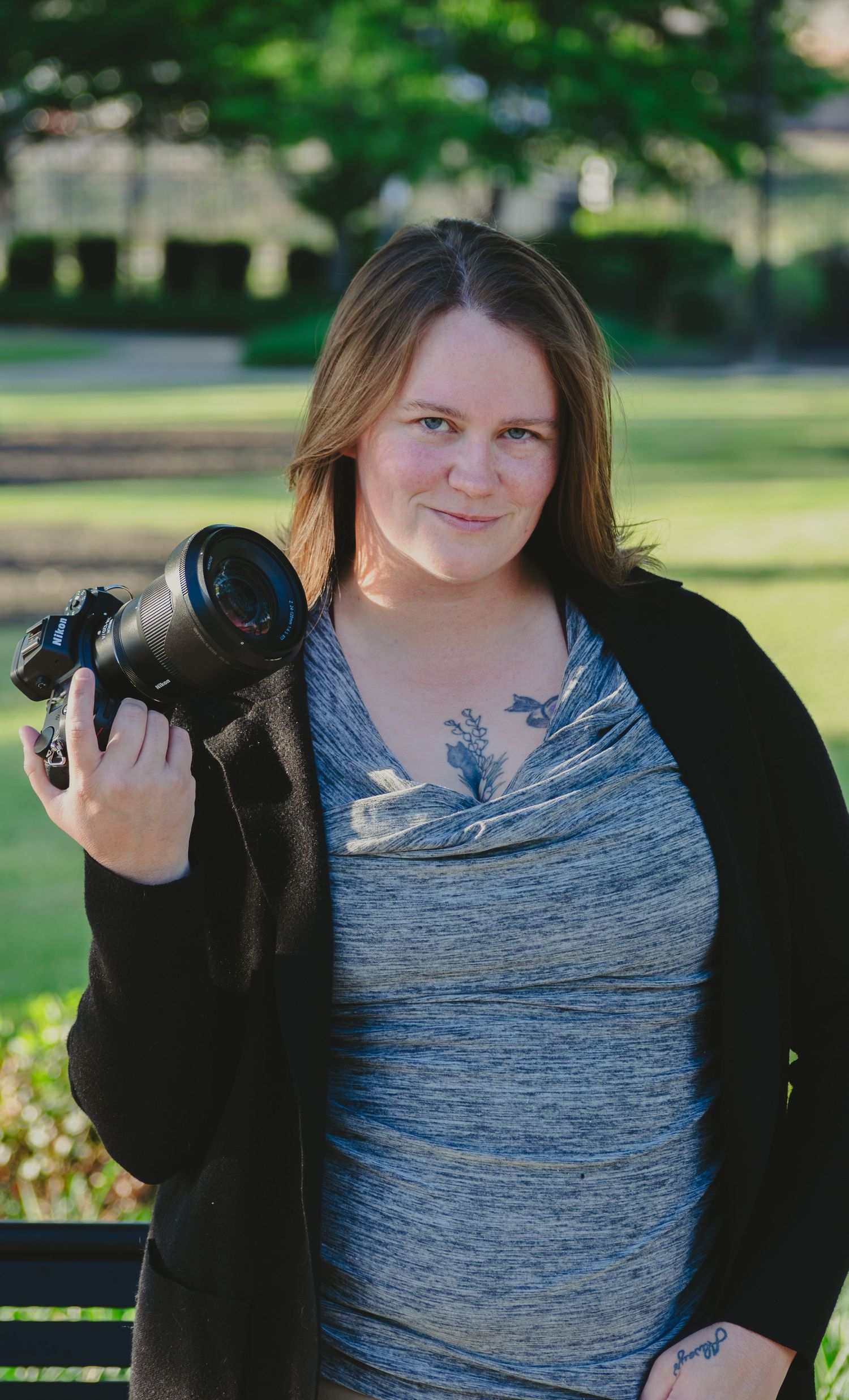 Female photographer smiling and holding a DSLR camera outdoors in a park with green trees in the background.