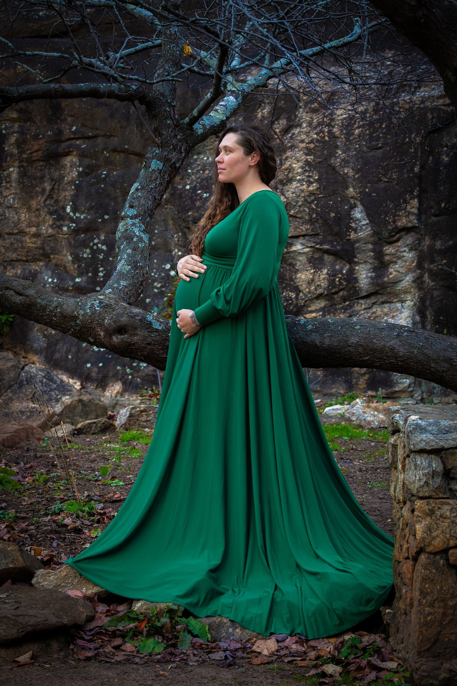 Pregnant woman in elegant green gown poses outdoors among rocks and bare trees in a serene maternity portrait.