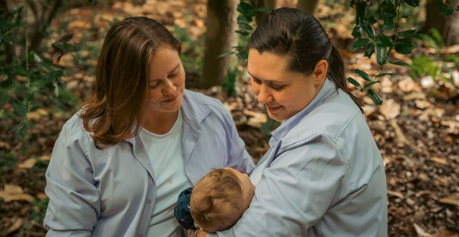 Two women in light blue shirts tenderly hold a newborn baby outdoors in a wooded setting.