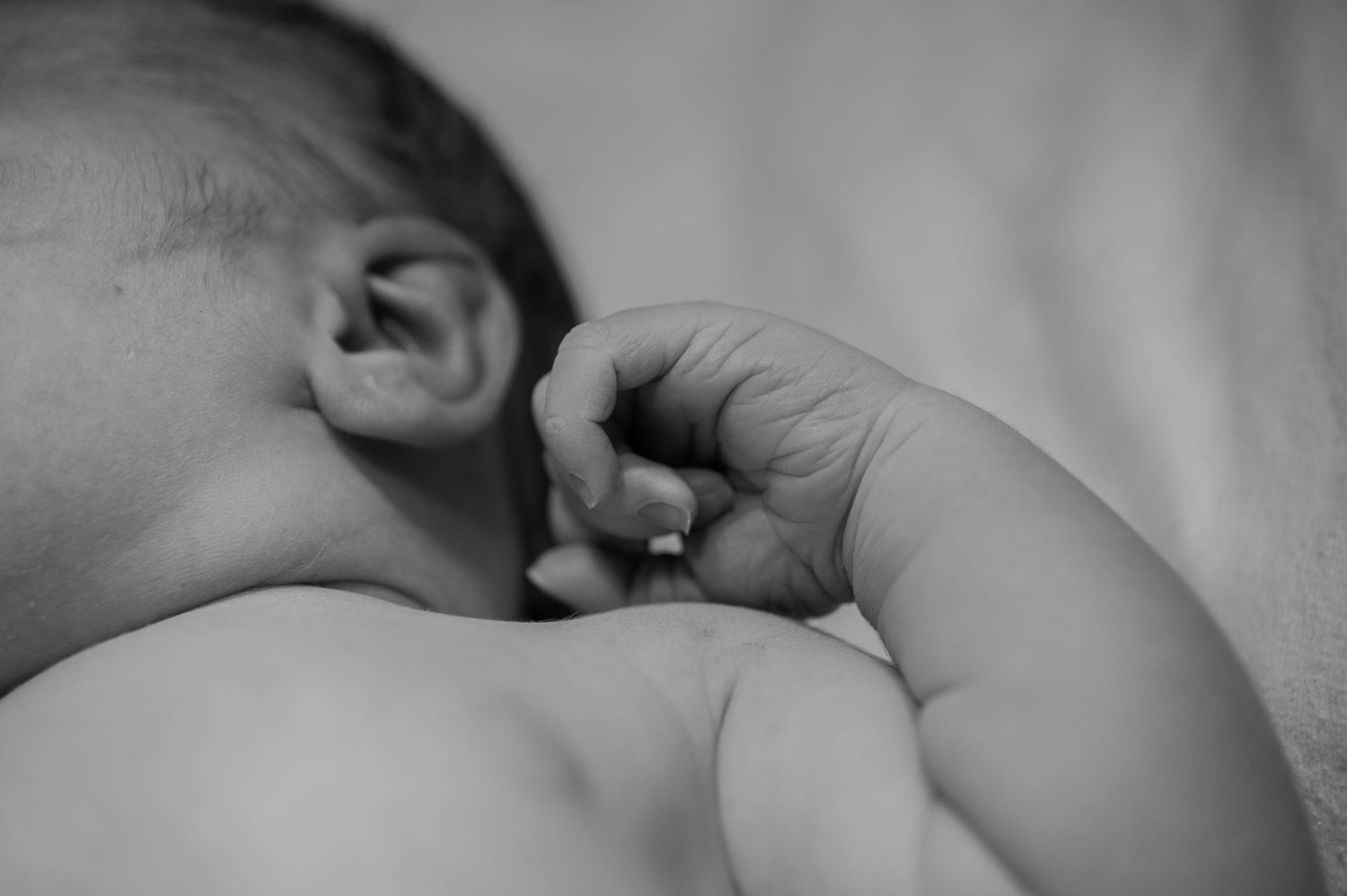 Close-up black and white photo of a sleeping newborn baby with tiny hand raised near face.