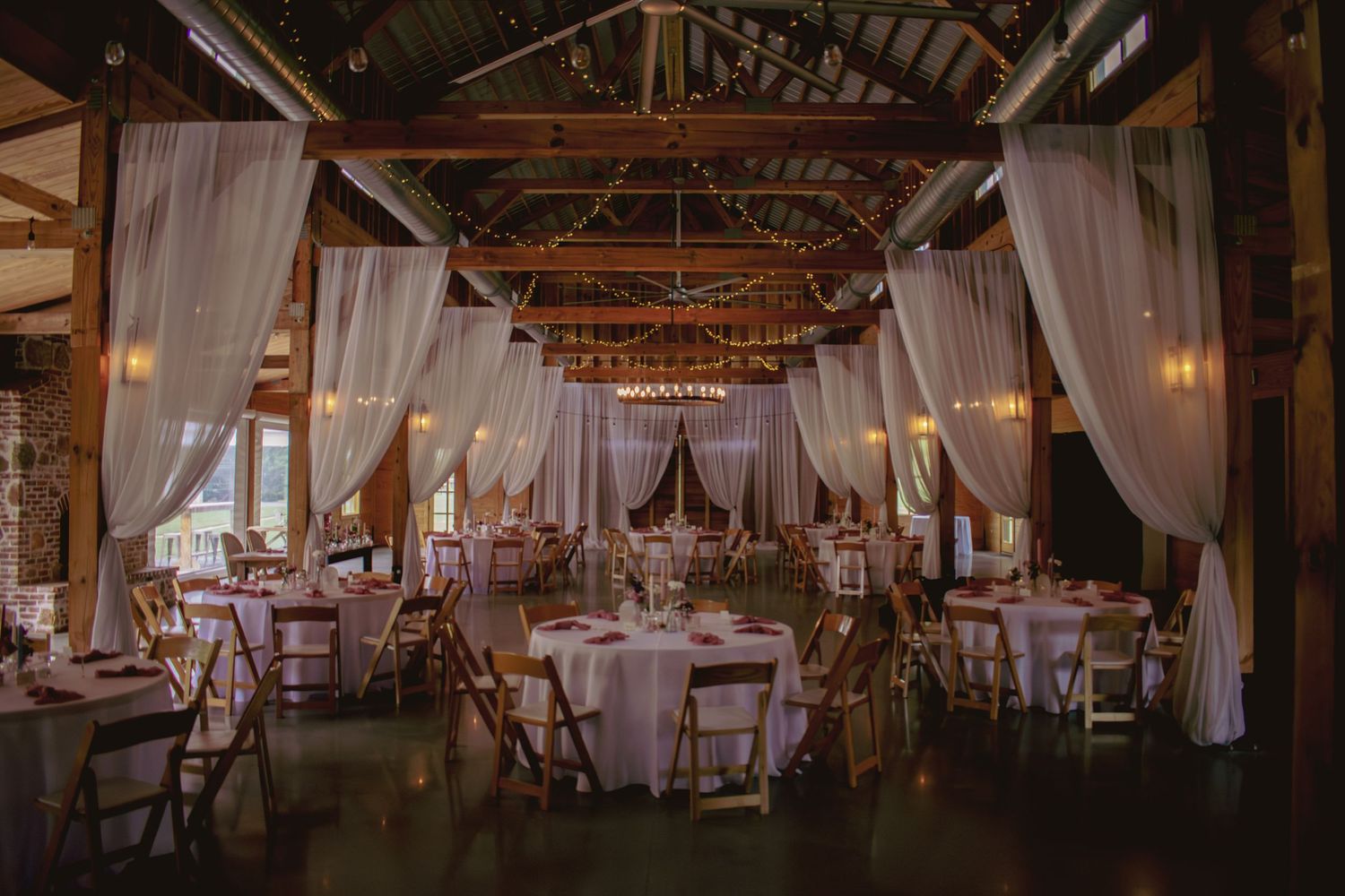 Elegant barn wedding reception with white draping, wooden beams, round tables, and warm ambient lighting.