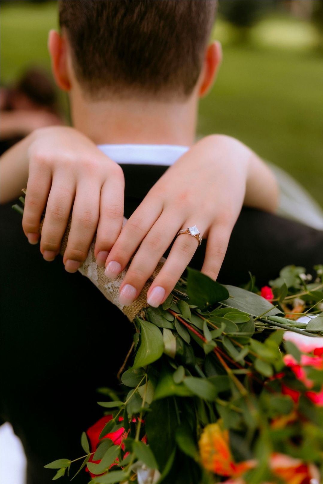 Bride with engagement ring hugging groom while holding colorful floral bouquet with greenery at outdoor wedding.