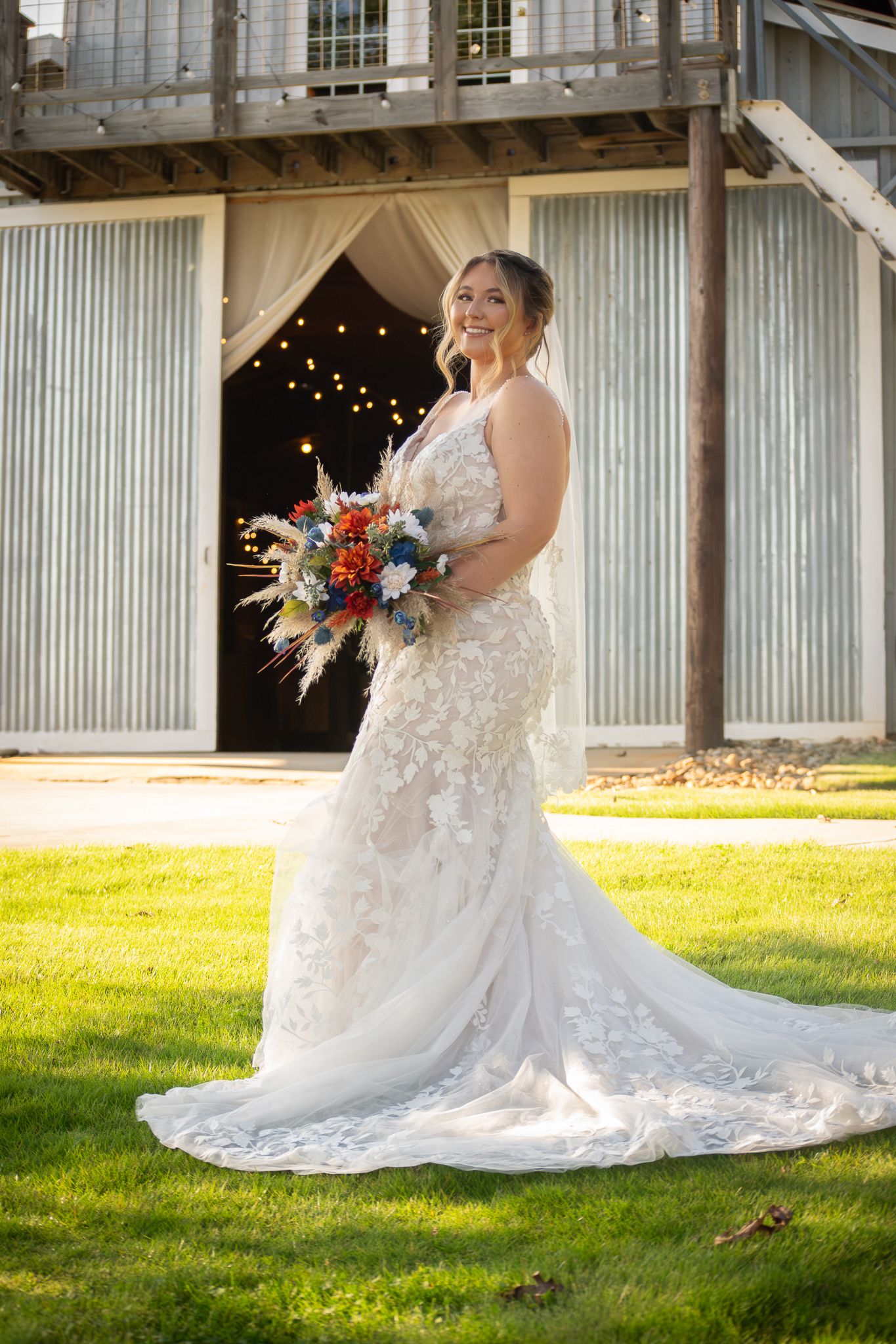 Bride in lace mermaid gown holding colorful bouquet stands on green lawn before rustic barn with string lights.