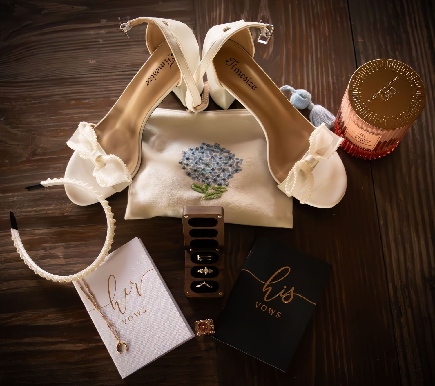A flat lay of elegant wedding accessories including bridal heels, vow books, rings, and a candle on wood.