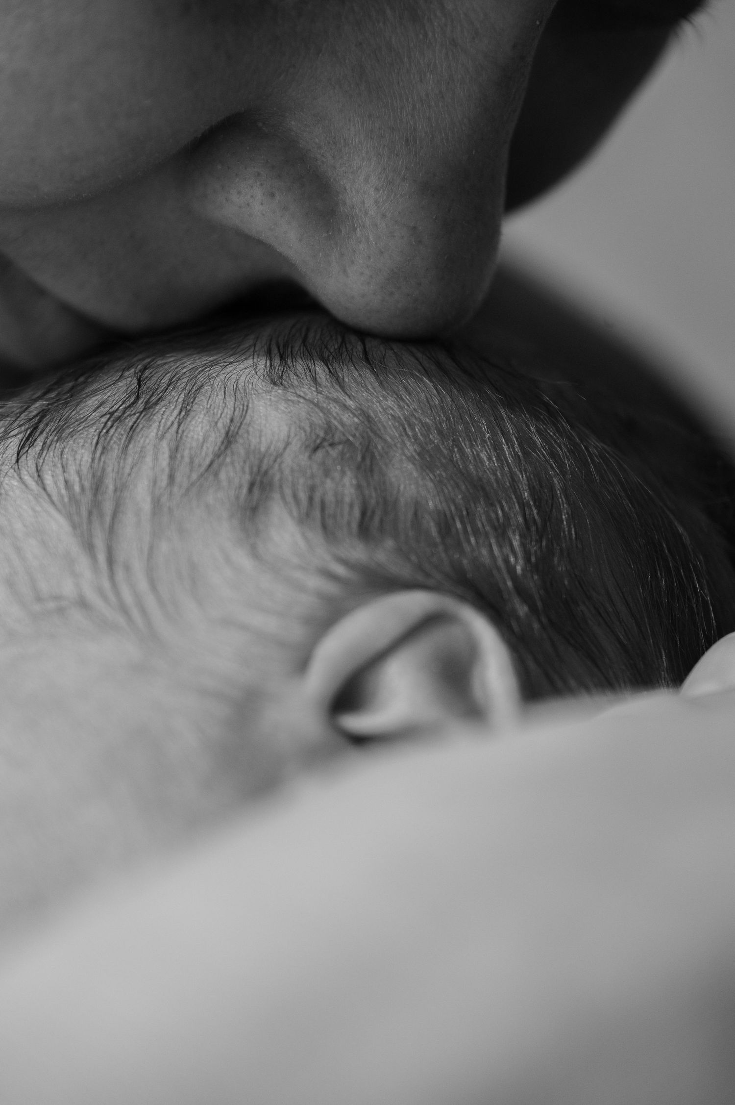 Black and white close-up of a parent gently kissing a sleeping newborn baby on the head.