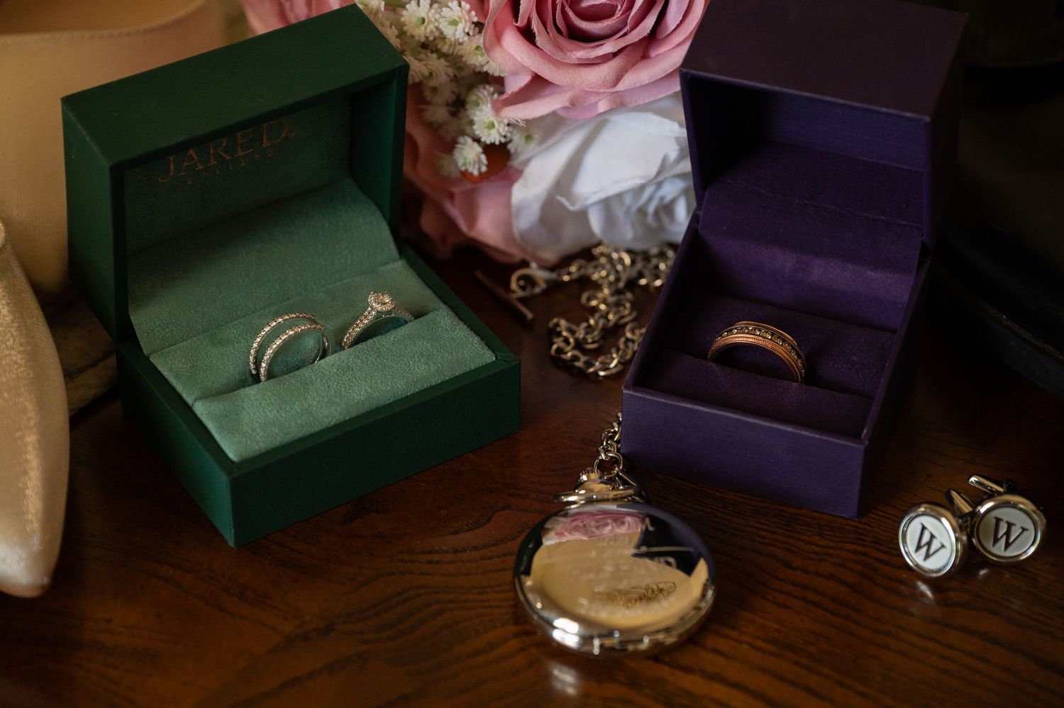 Wedding rings in green and purple boxes with a vintage pocket watch and monogram cufflinks on a wooden surface.