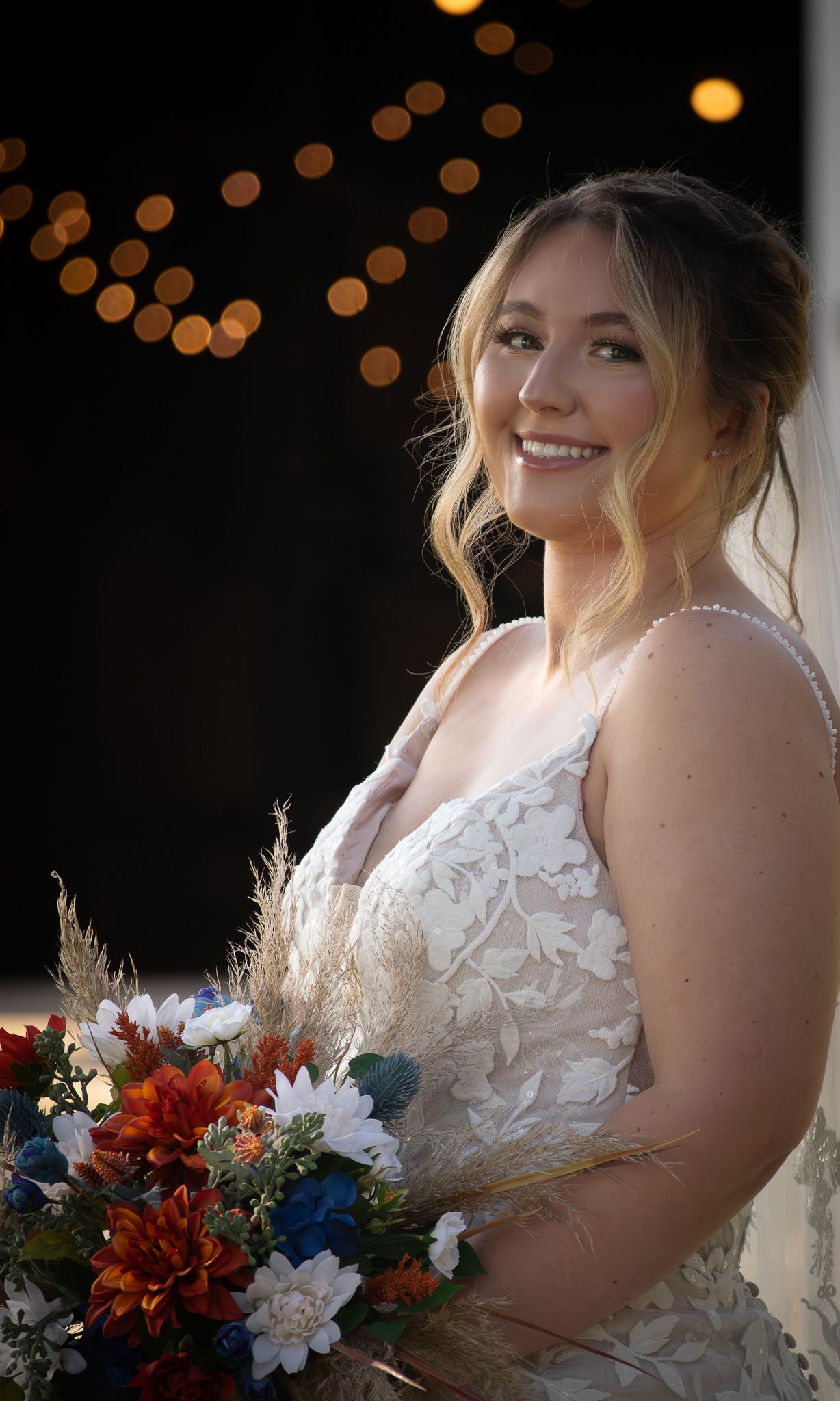 Smiling bride in lace wedding dress holding colorful bouquet with bokeh lights in background.