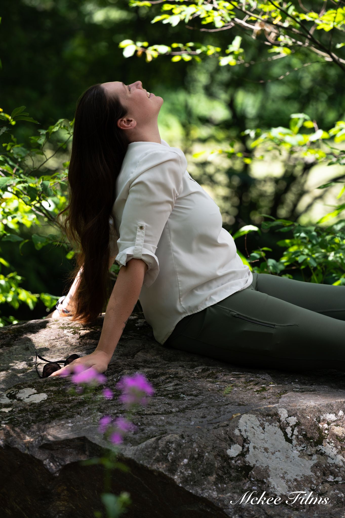 Pregnant woman in white shirt reclining on rock surrounded by green forest foliage.