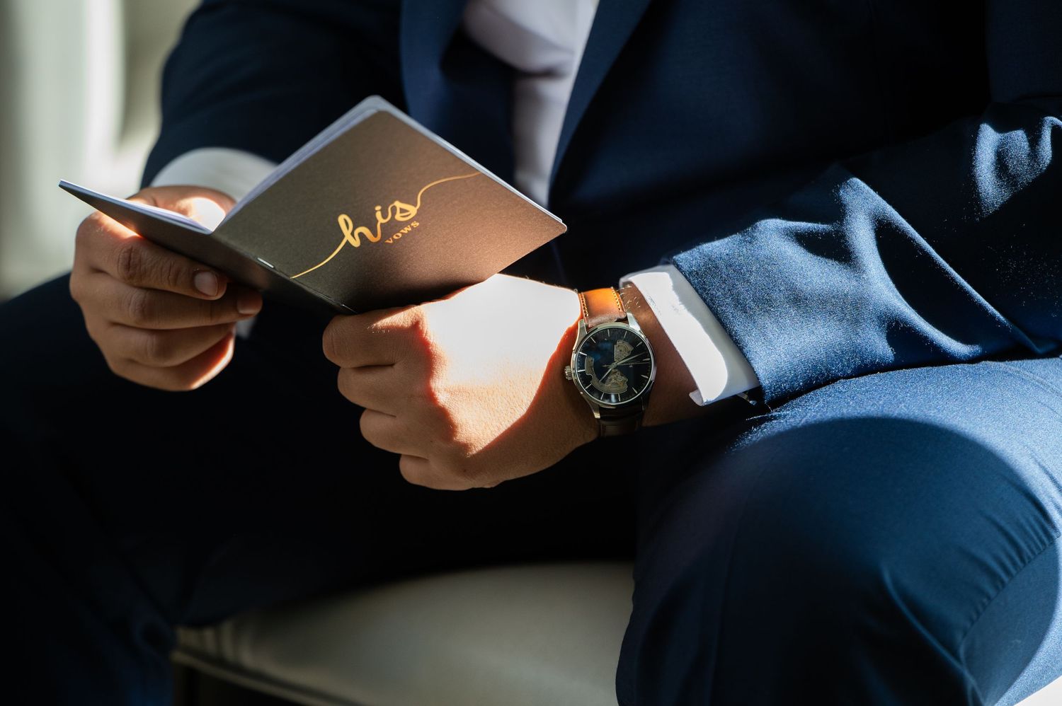 Man in navy suit holding a 'his' wedding card, wearing a luxury watch, elegant groom preparation detail.