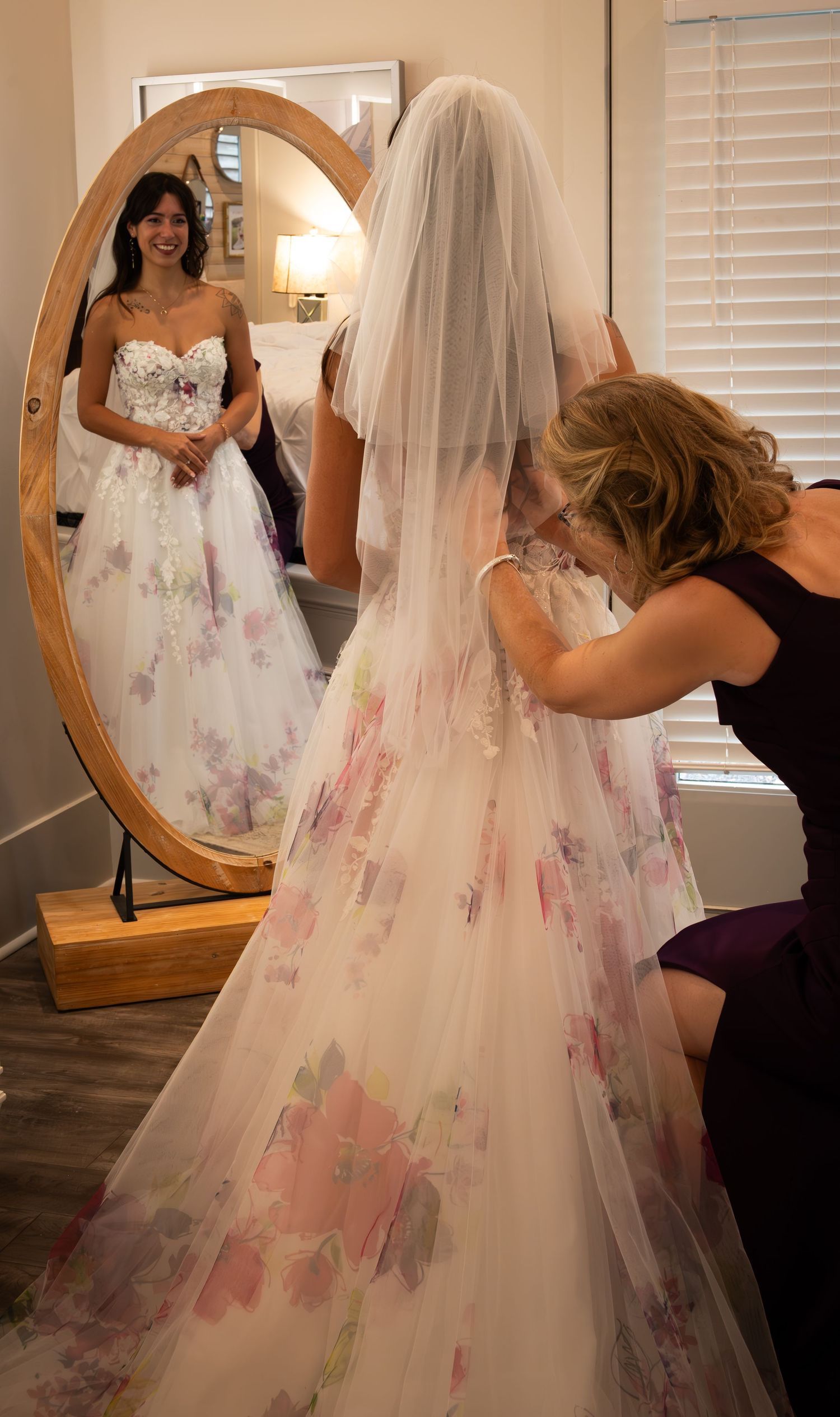 A bride in a floral wedding gown and veil smiles at her reflection in an oval mirror while an attendant adjusts her veil.