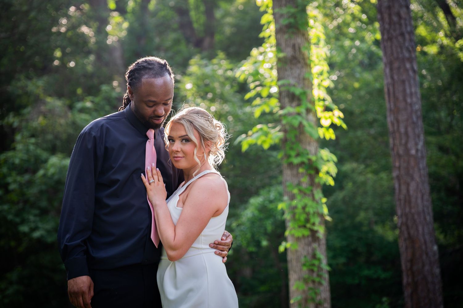 Couple in formal attire embracing outdoors, groom in black shirt with pink tie, bride in white dress, lush green forest backdrop.