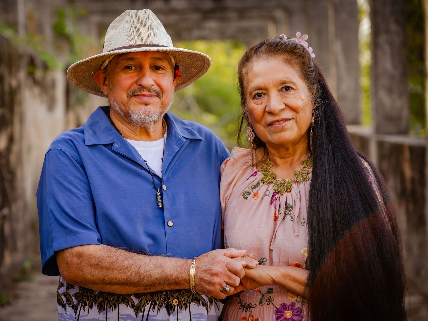 An elderly Hispanic couple smiles warmly while holding hands outdoors, the man wearing a white hat and blue shirt.