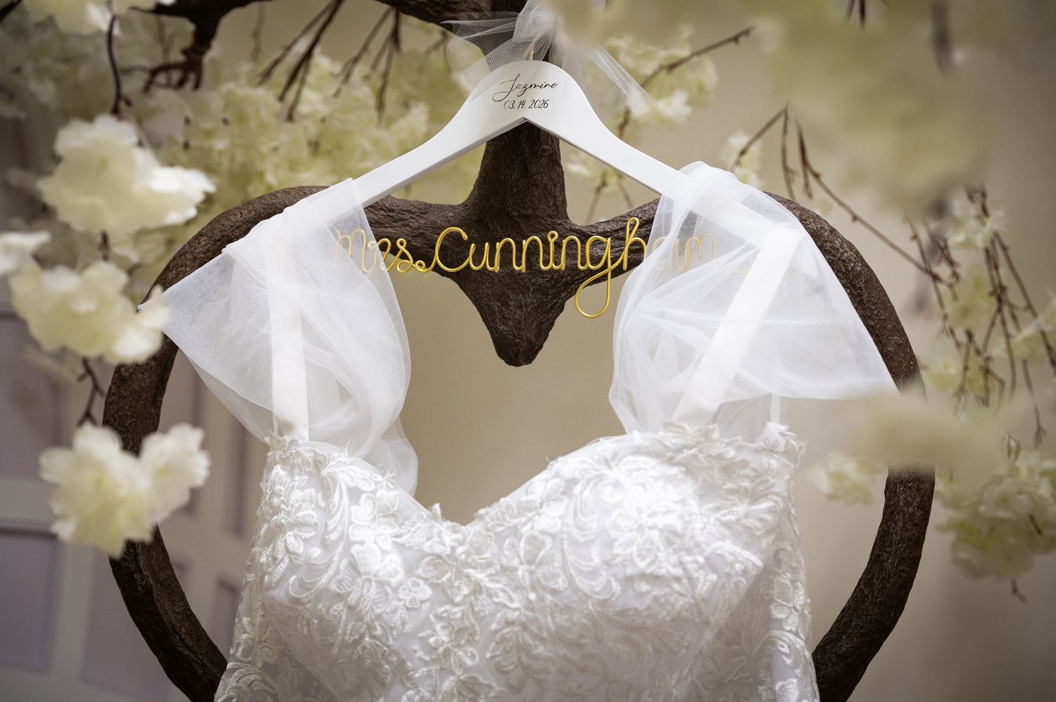 White lace wedding dress hanging on a personalized Mrs. Cunningham hanger surrounded by white floral decor.
