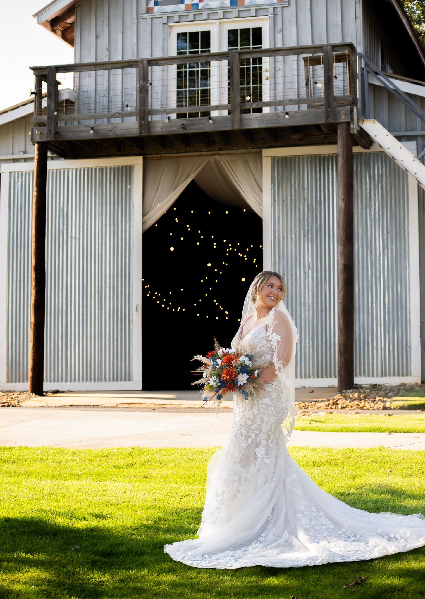 Bride in lace wedding gown holding colorful bouquet stands before rustic barn venue with string lights.