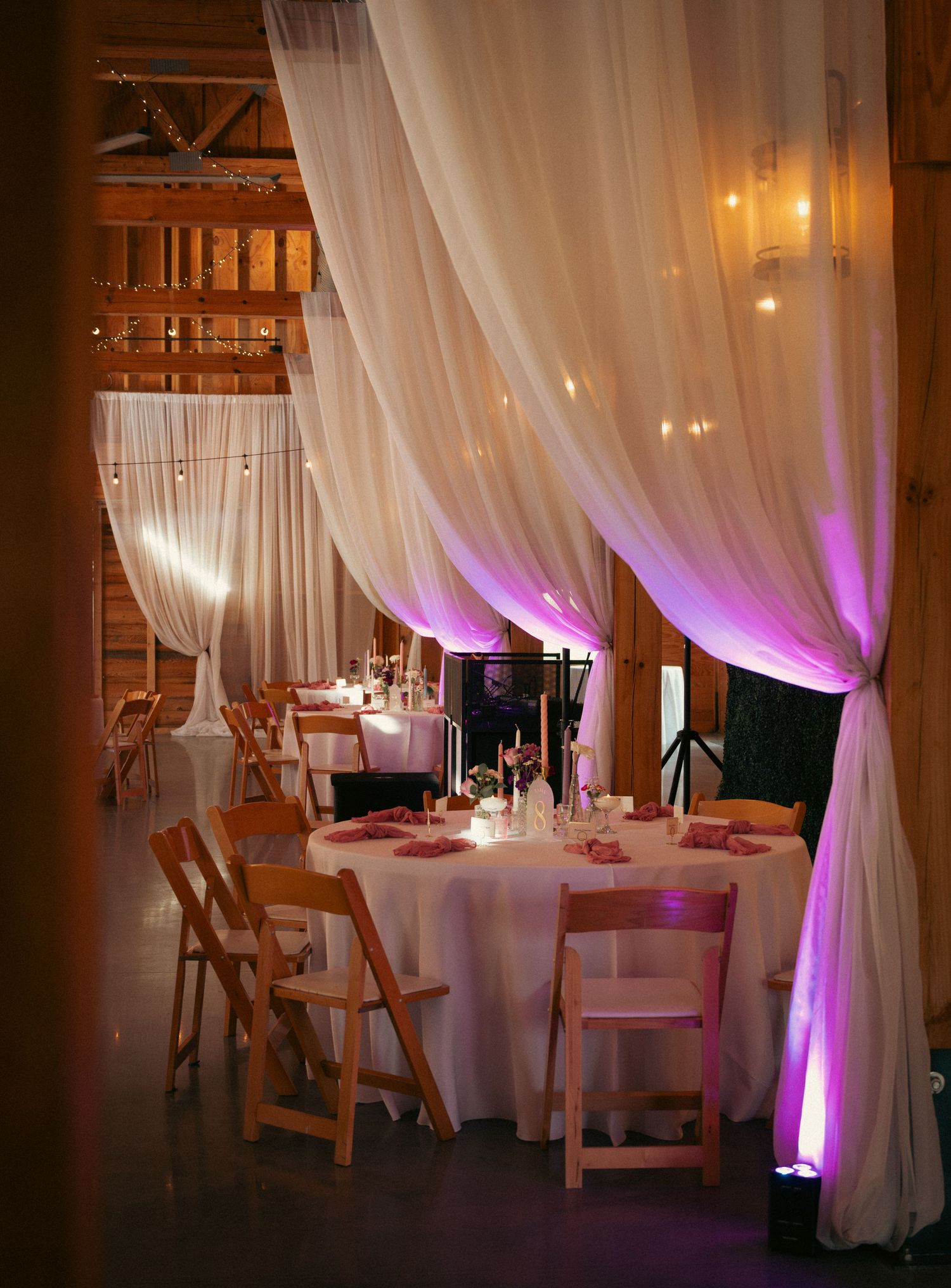 Elegant barn wedding reception with white draping, string lights, round tables, wooden chairs, and purple uplighting.