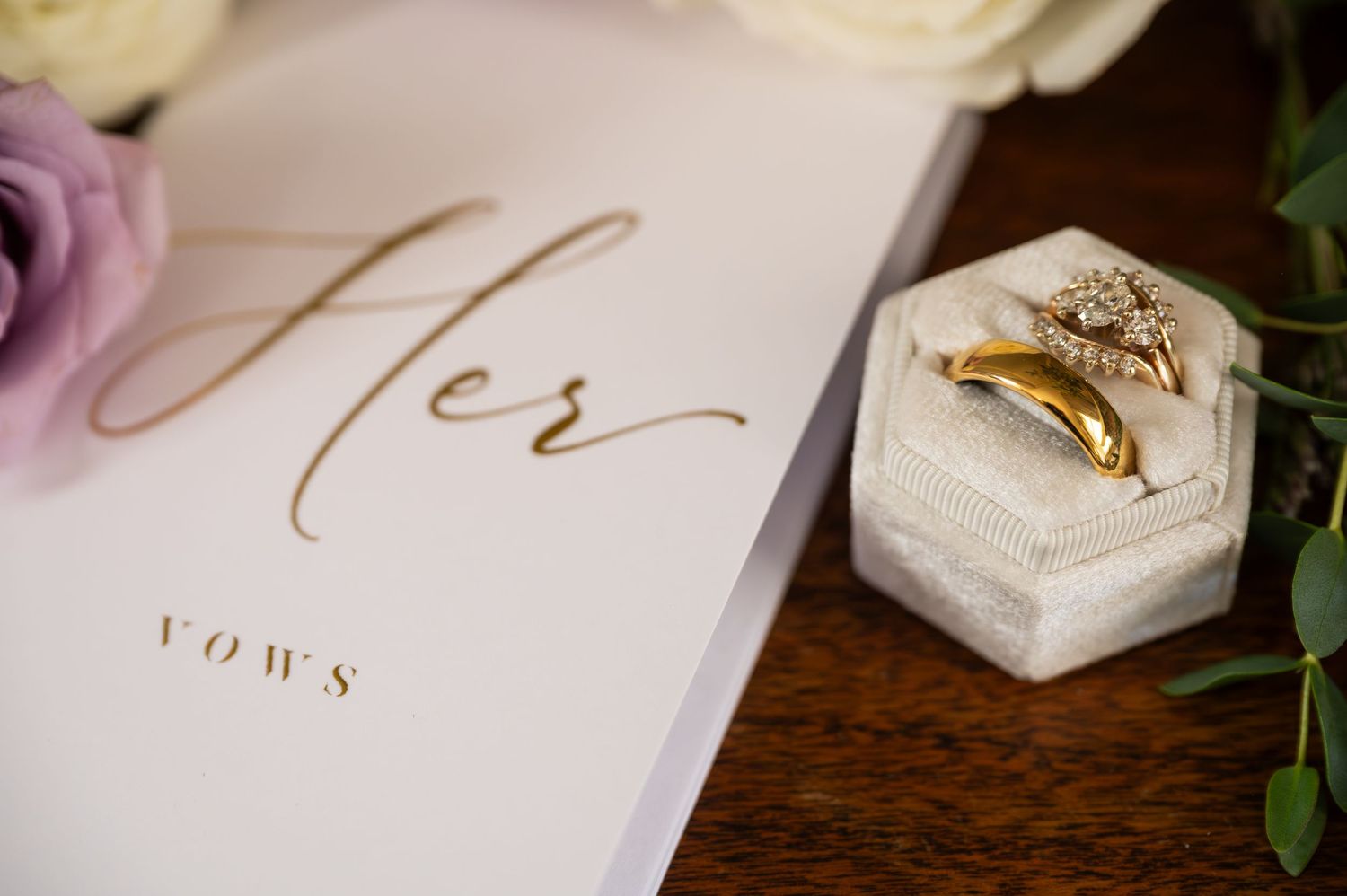 Gold wedding rings in a velvet hexagon box beside a white Her Vows book, surrounded by soft purple and white roses.
