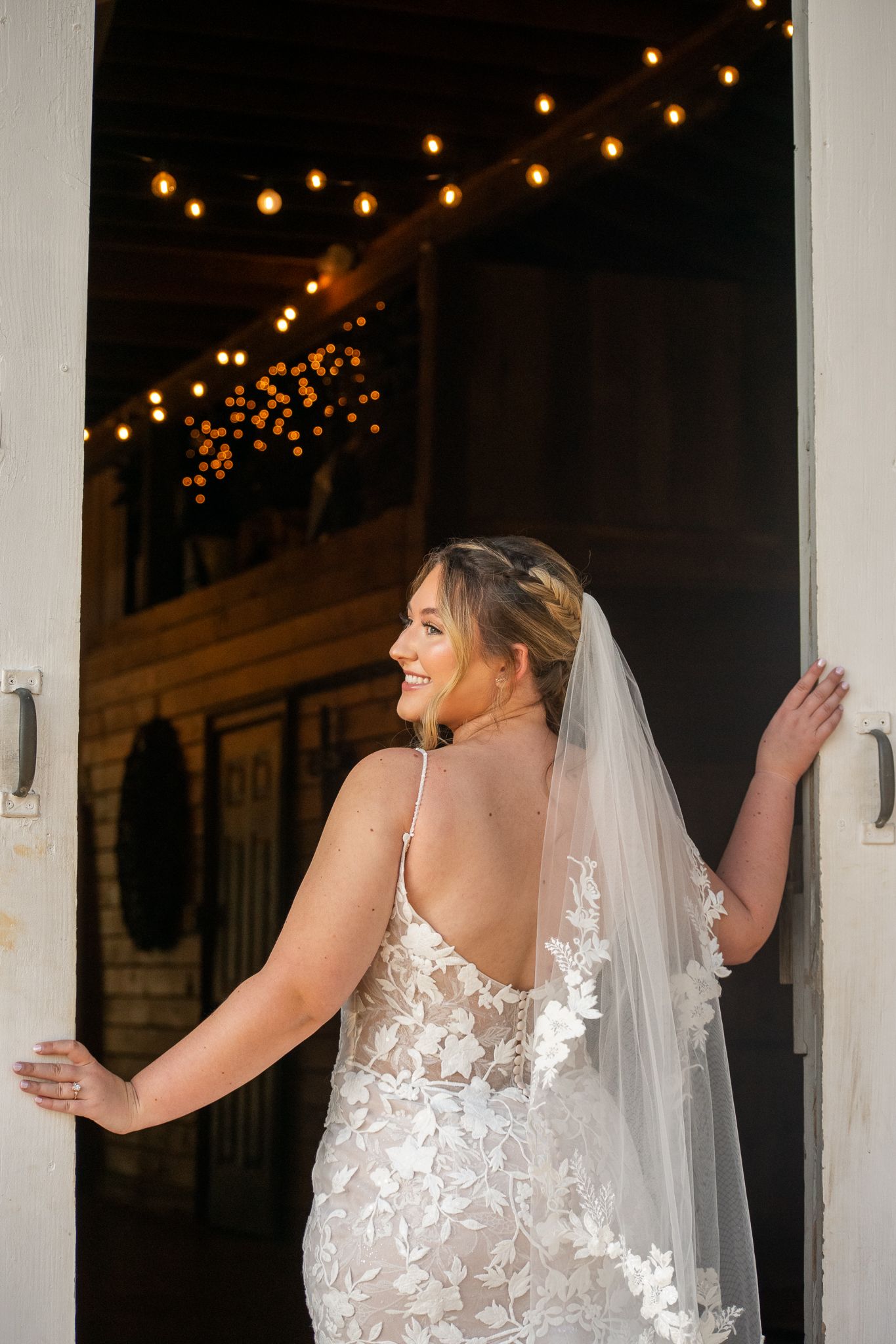 Bride in lace backless wedding gown and veil stands in barn doorway with warm string lights glowing behind her.