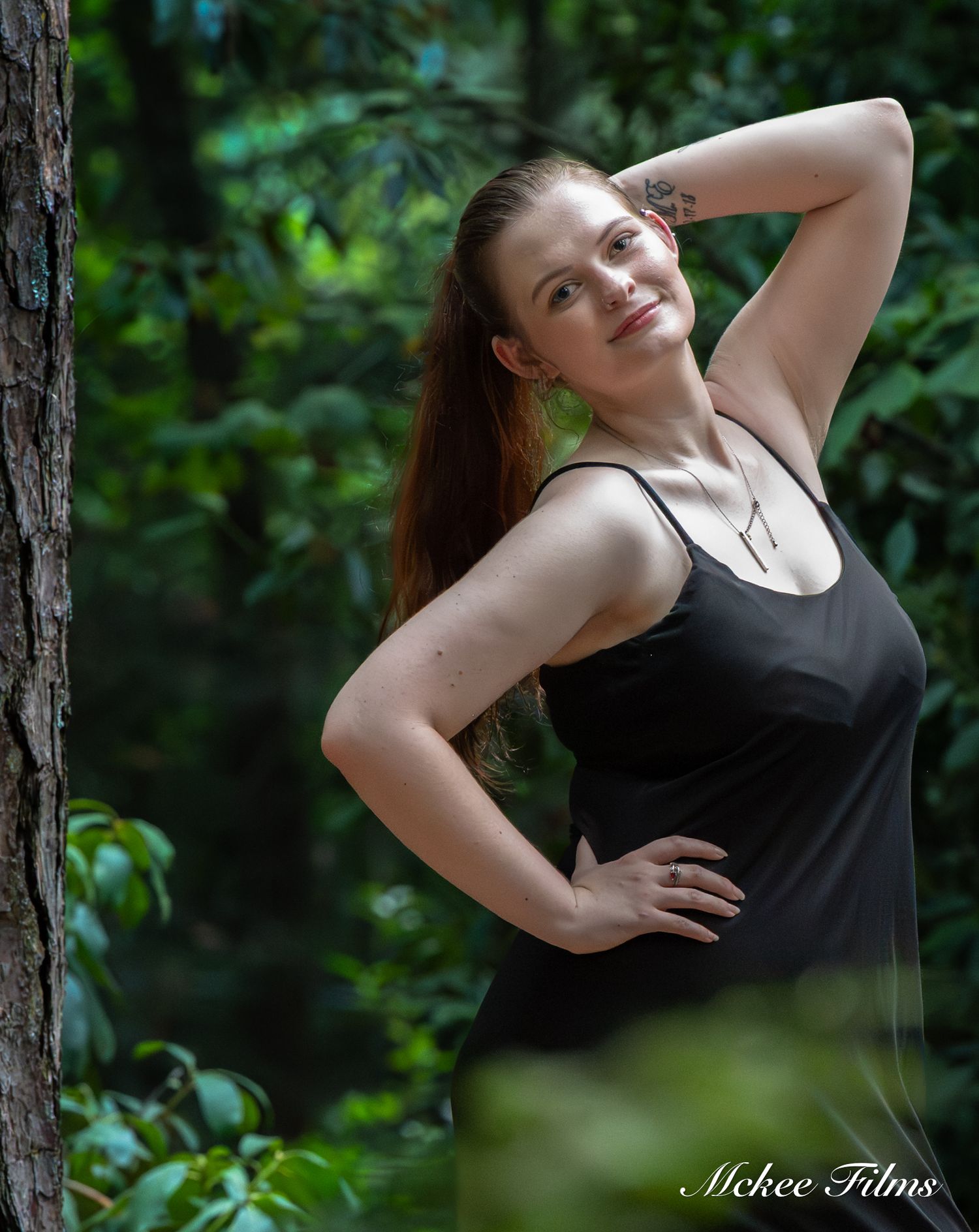 A smiling woman in a black dress poses gracefully among lush green forest trees in a professional outdoor photo shoot.