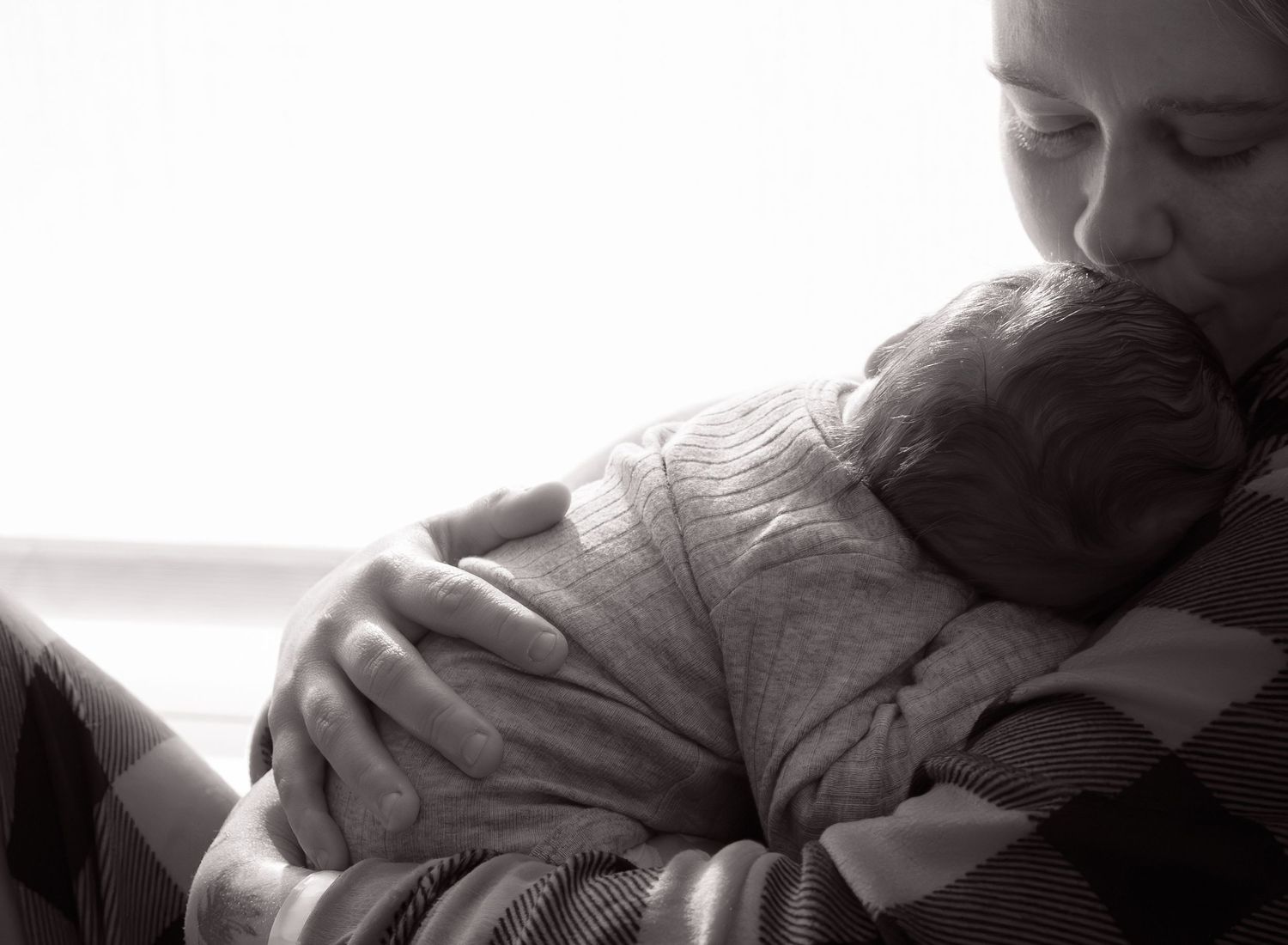 Black and white photo of a mother tenderly kissing her newborn baby while cradling the infant in her arms.