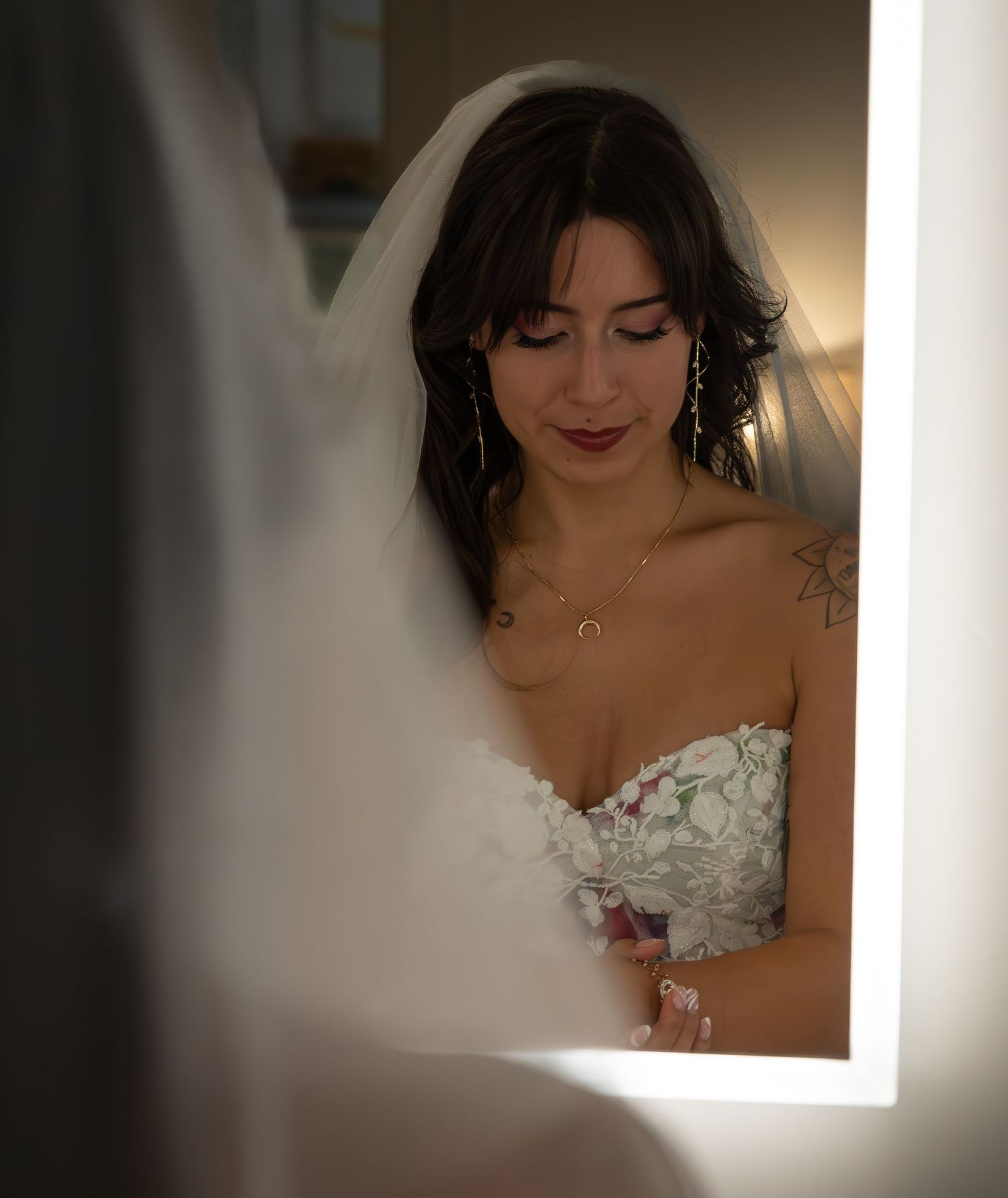 Bride in strapless floral gown and veil gazes down at bouquet, reflected in a lit vanity mirror.