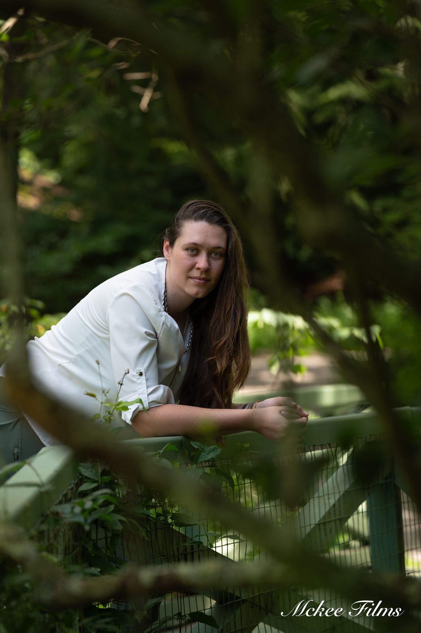 Young woman in white shirt leaning on a green fence surrounded by lush green trees in a nature setting.