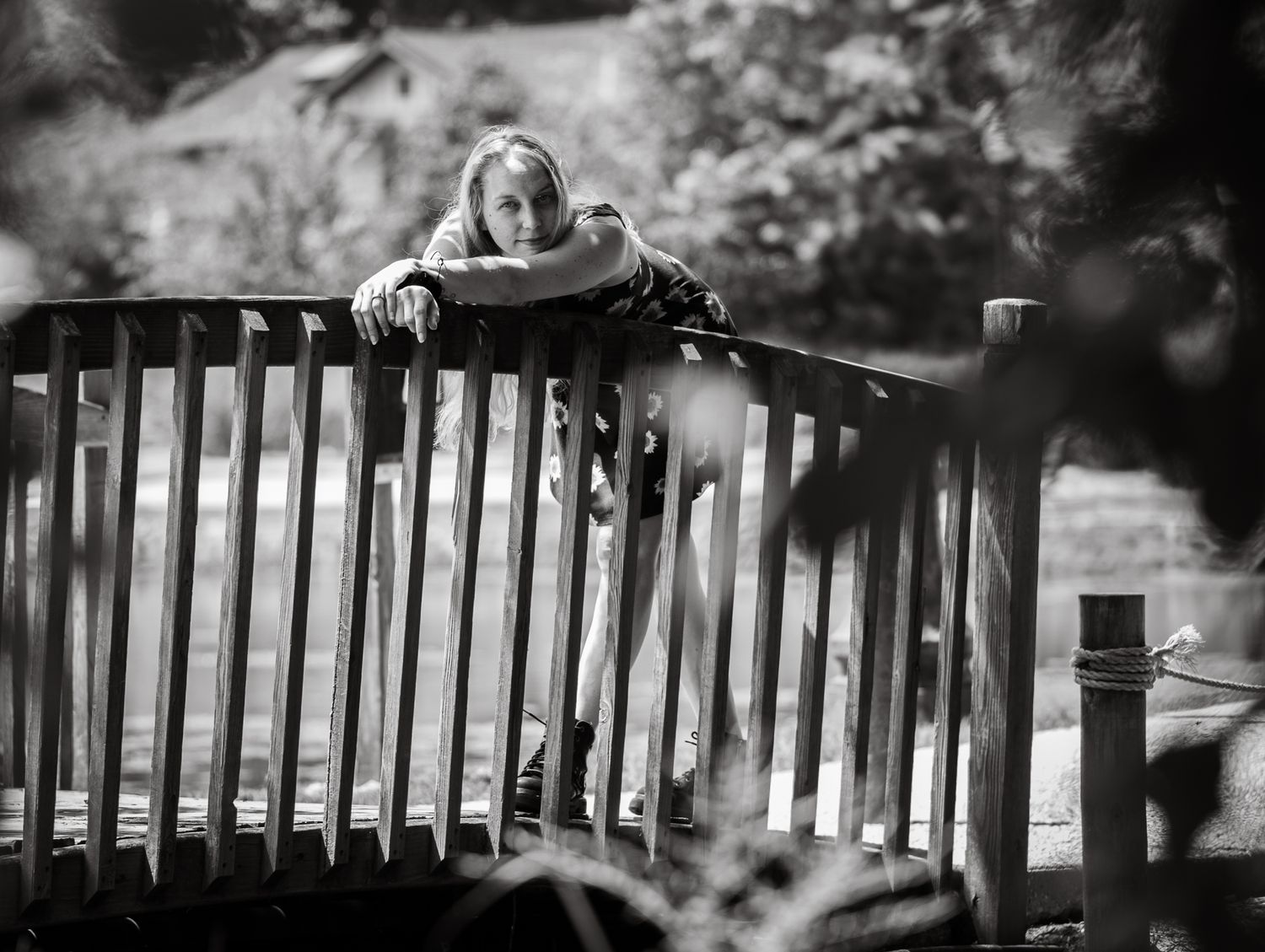 Black and white photo of a woman leaning over a wooden park bench, surrounded by blurred greenery.
