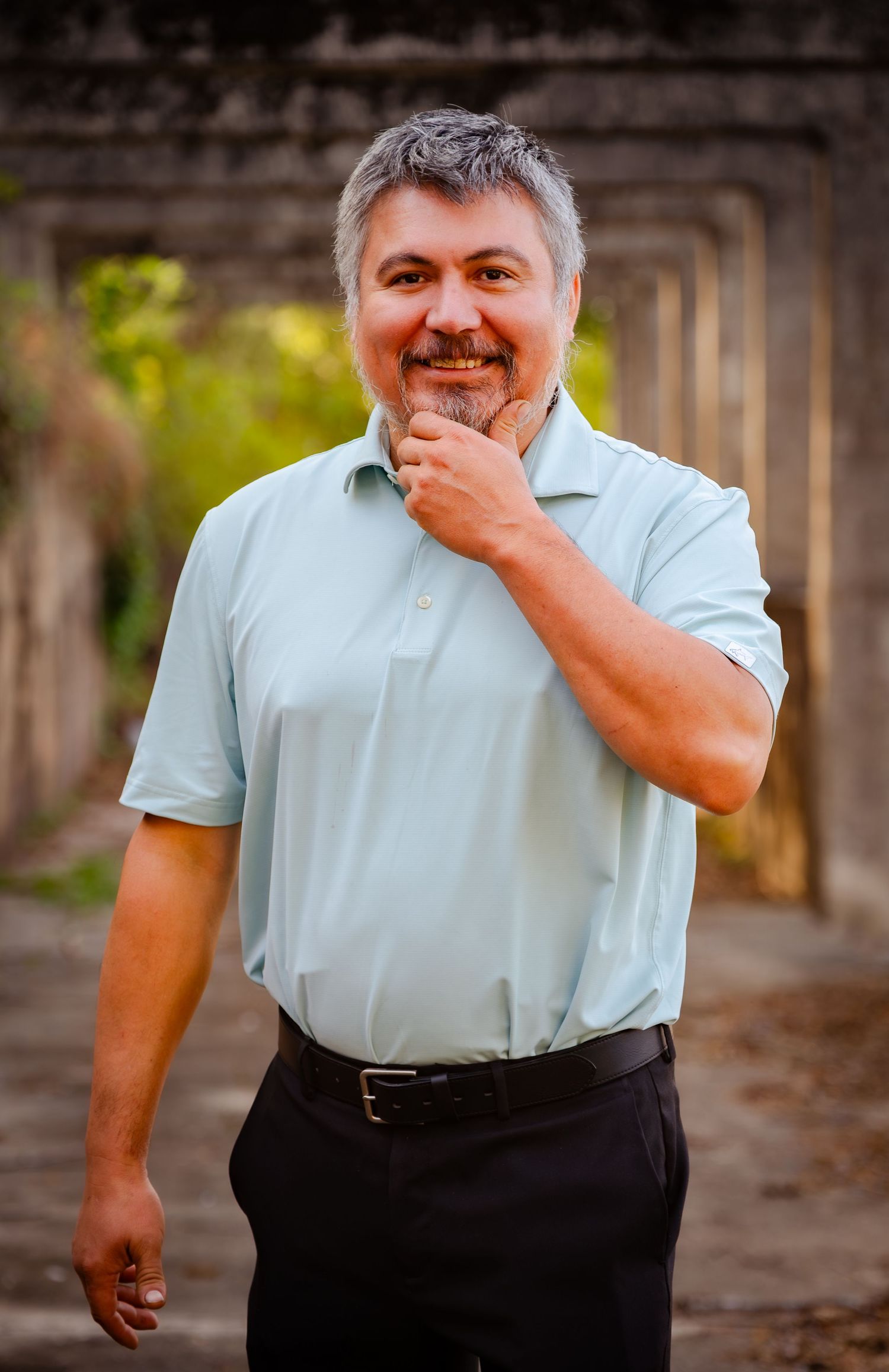 Smiling middle-aged man with gray hair in a light blue polo shirt posing outdoors near stone archway.