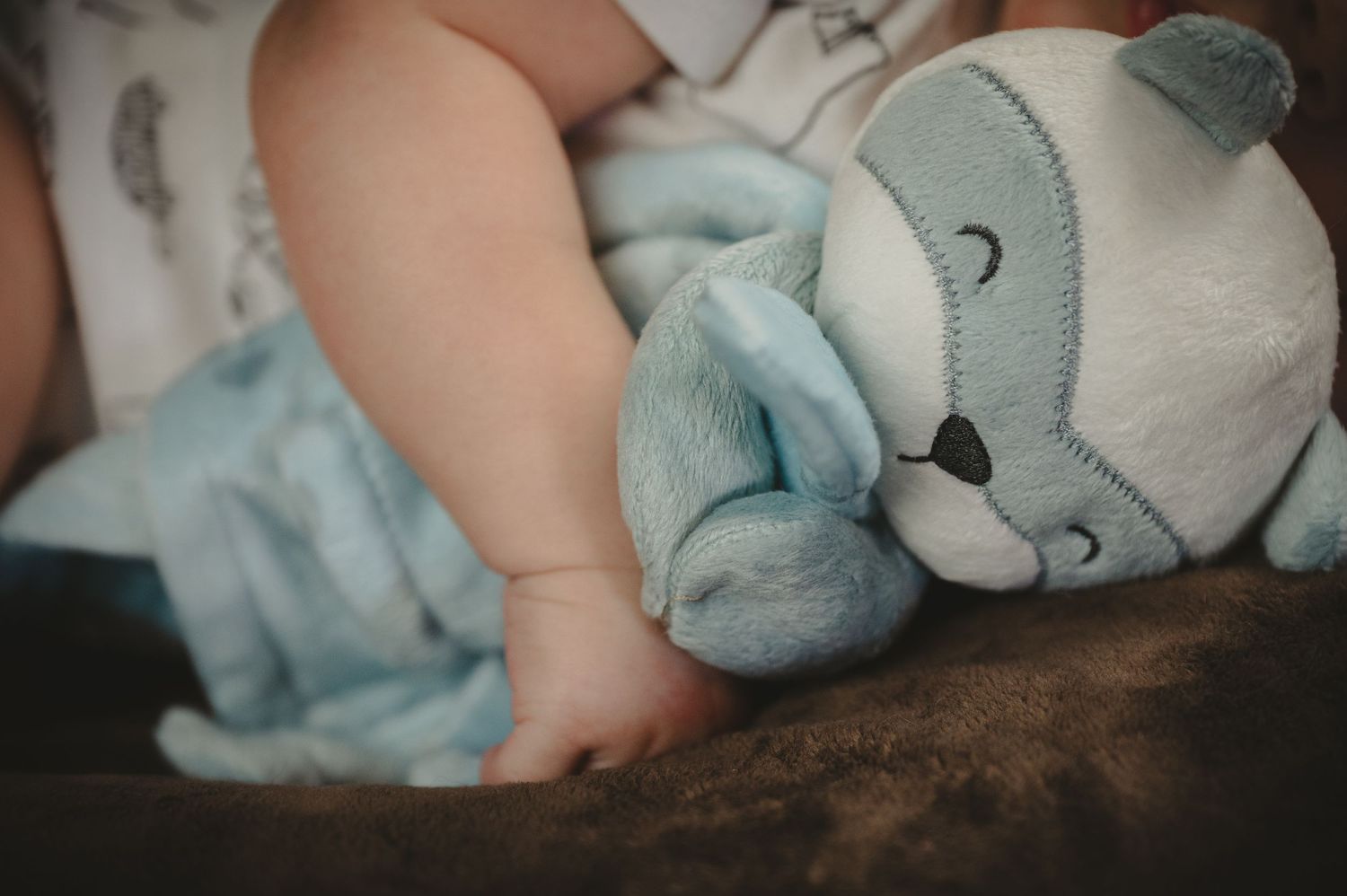 A sleeping baby cuddles a soft blue teddy bear plush toy on a brown surface.