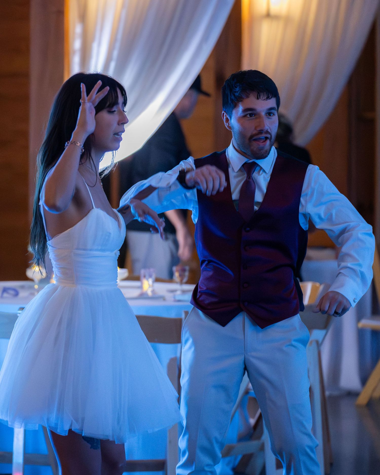 Bride and groom dancing together at wedding reception, wearing white dress and burgundy vest with white pants.