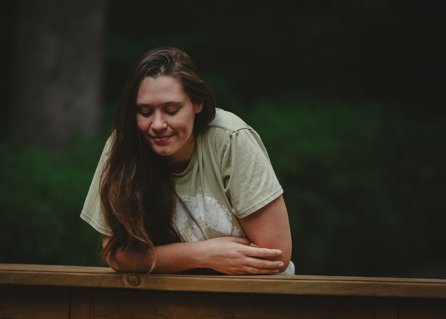 Young woman with long hair smiles warmly while leaning on a wooden railing outdoors.