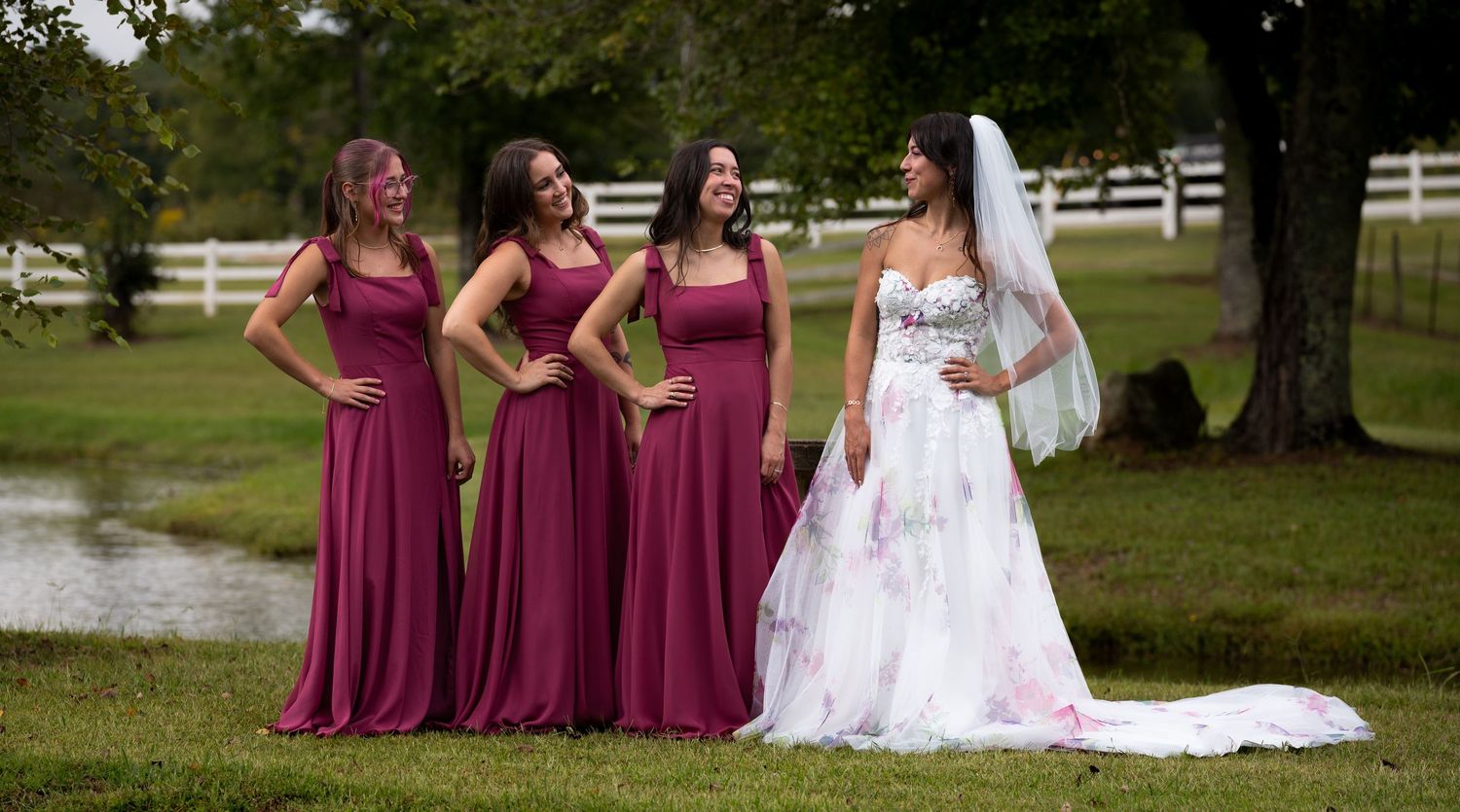 Bride in floral wedding dress laughs with three bridesmaids wearing matching burgundy gowns outdoors.