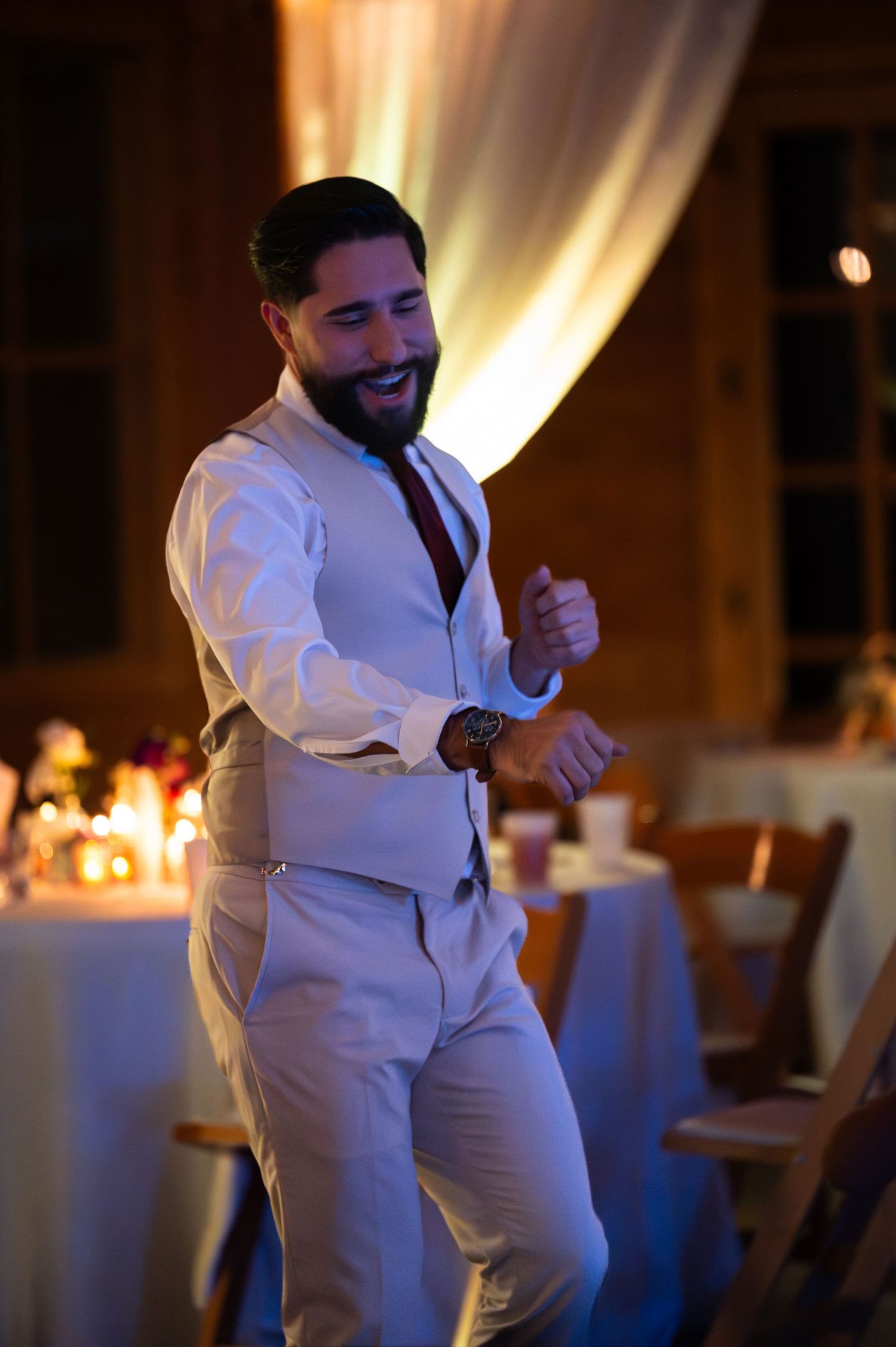 A bearded groom in a white vest suit dances joyfully at a candlelit wedding reception.