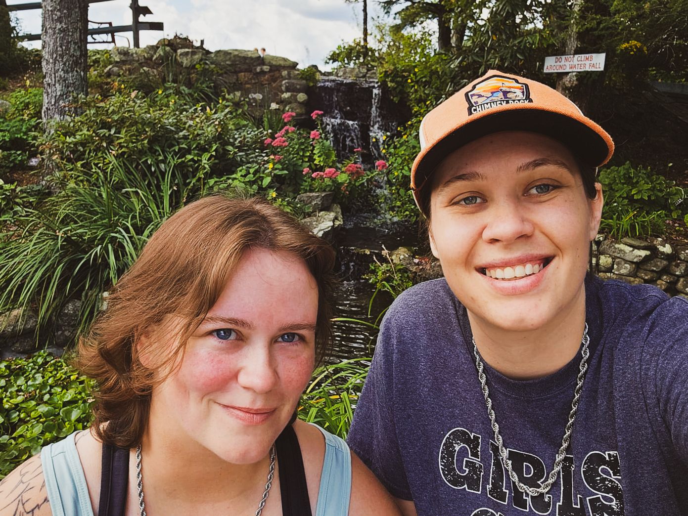 Two young women smiling together in a selfie outdoors near a lush garden with a waterfall backdrop.