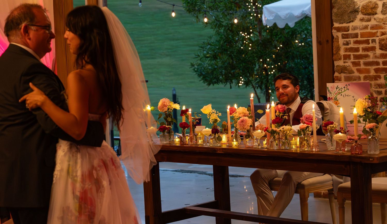 Bride and groom dancing at candlelit wedding reception with floral centerpieces and string lights in rustic venue.