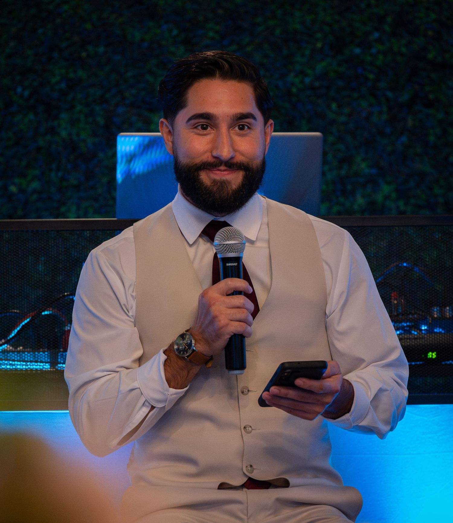 Smiling bearded man in white vest and tie holding microphone and phone at formal event.