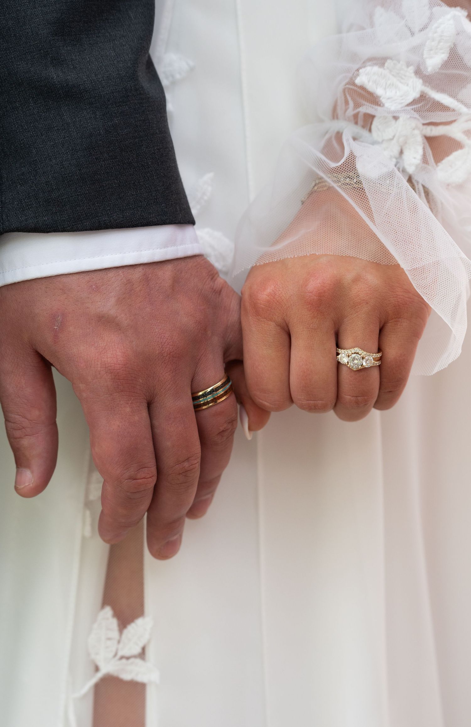 Close-up of newlywed couple holding hands, showcasing wedding rings on ring fingers, bride in white lace gown.