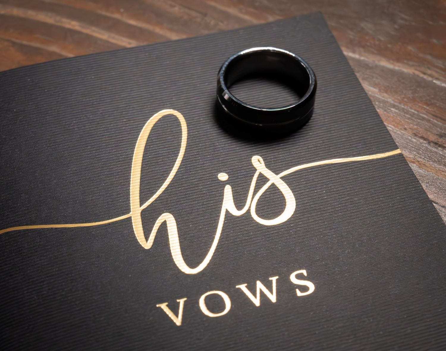 Black wedding vow book with gold script reading his vows, topped with a classic black tungsten ring.