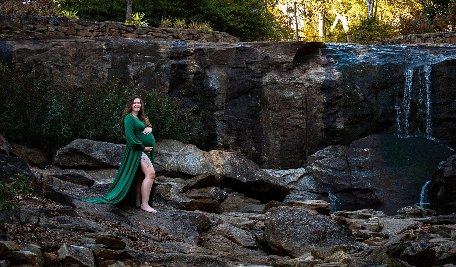 A pregnant woman in a flowing green gown poses gracefully on rocks near a scenic waterfall at sunset.