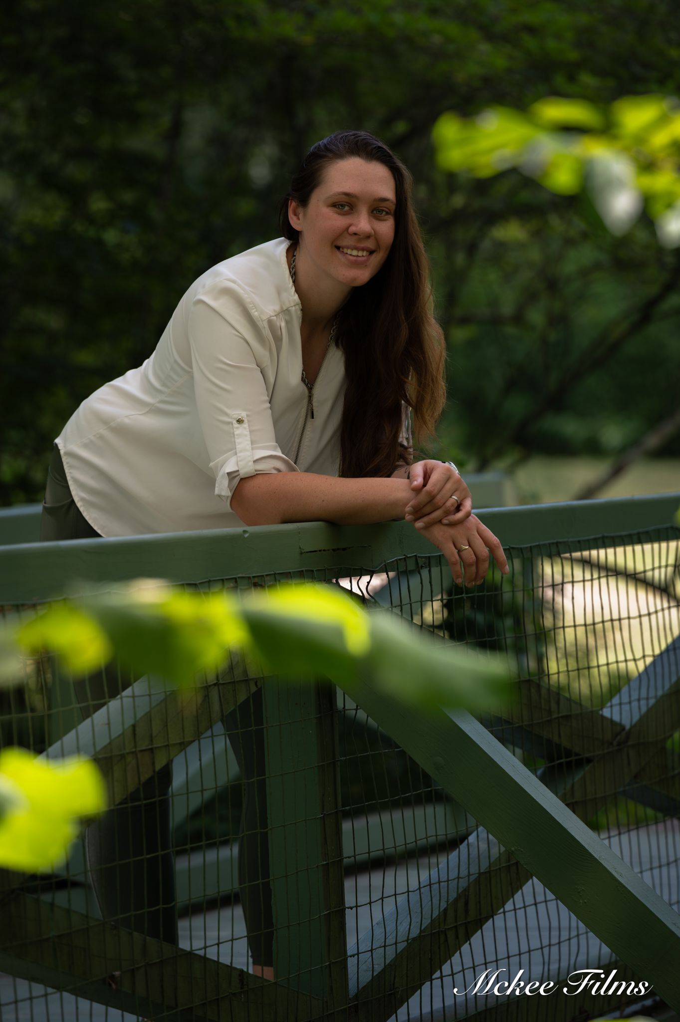 A smiling young woman with long brown hair leans on a green metal bridge railing surrounded by lush greenery.