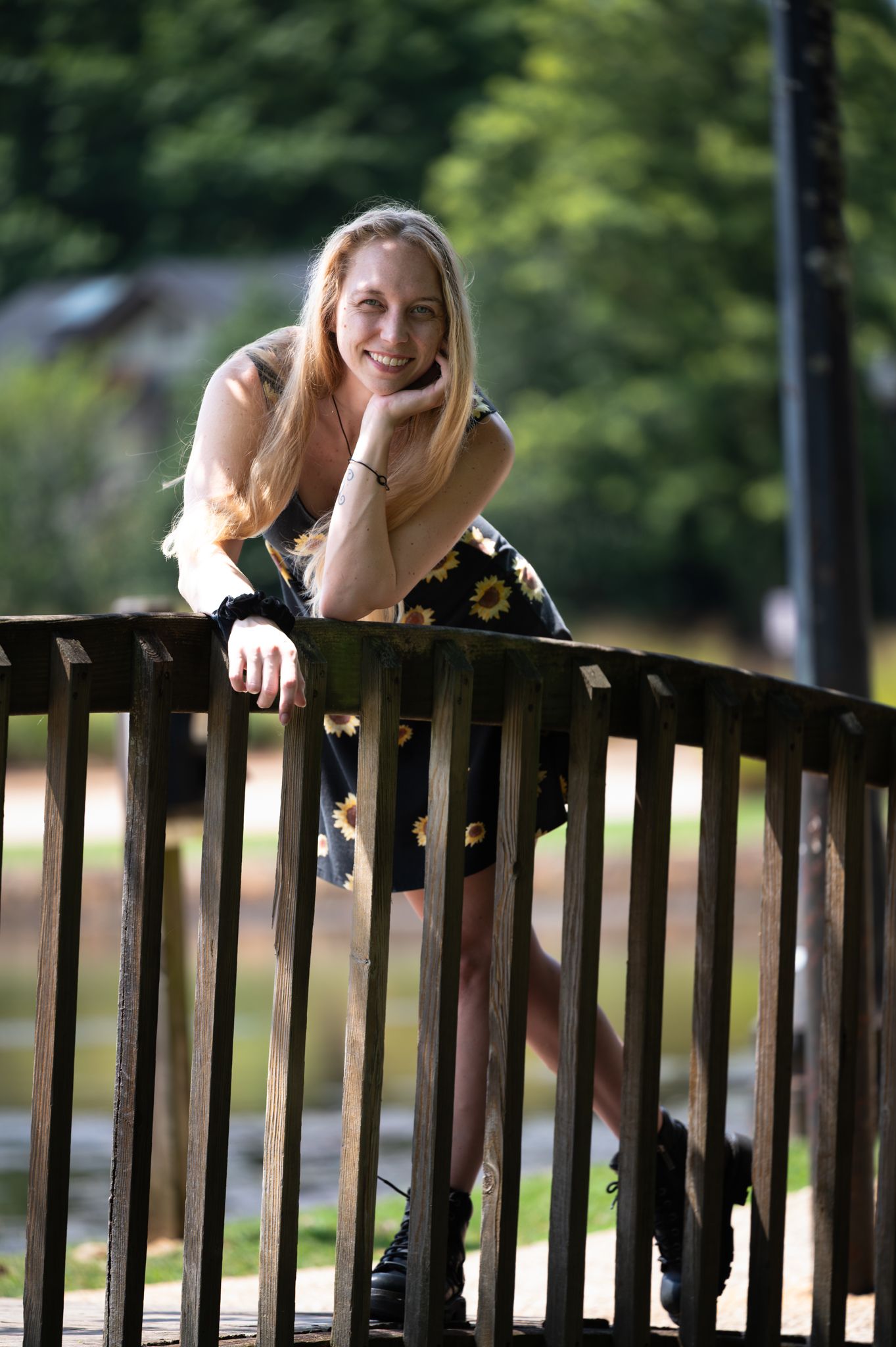 Smiling blonde woman in floral dress leaning on wooden railing outdoors with green trees in background.