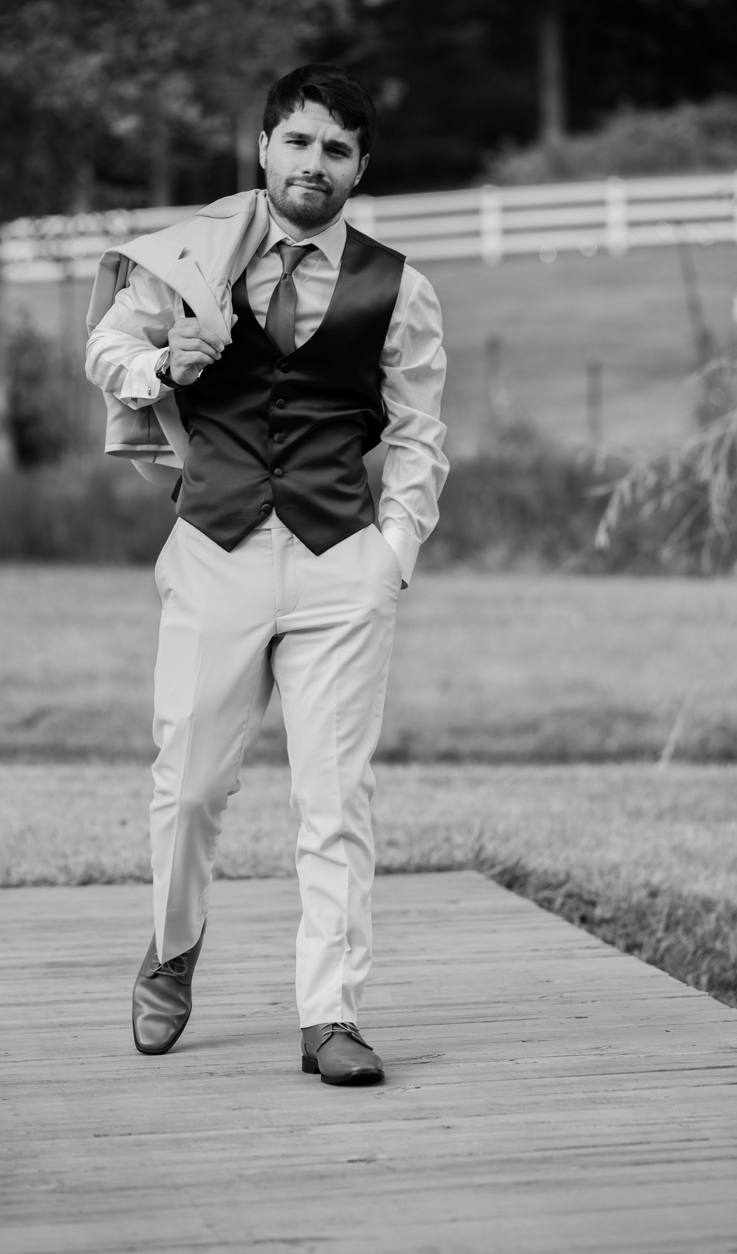 Black and white photo of groom carrying jacket over shoulder on wooden path.