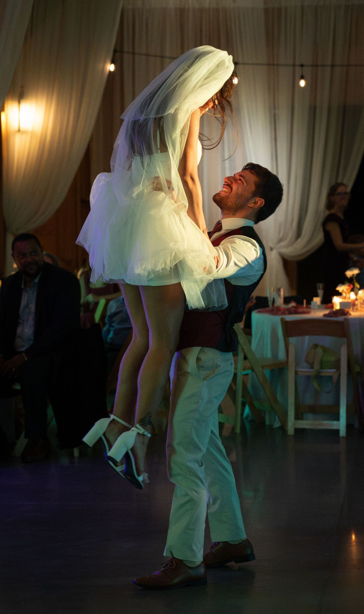 Groom joyfully lifts bride in white dress and veil during first dance at elegant wedding reception.