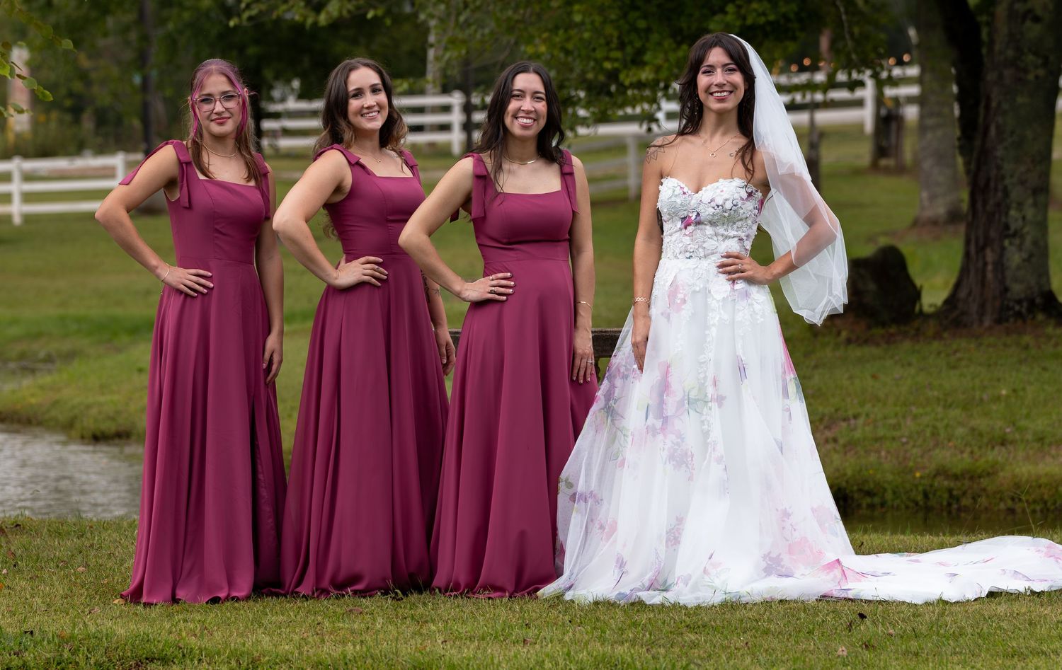 Bride in floral ball gown poses with three bridesmaids in burgundy dresses on lush green lawn.