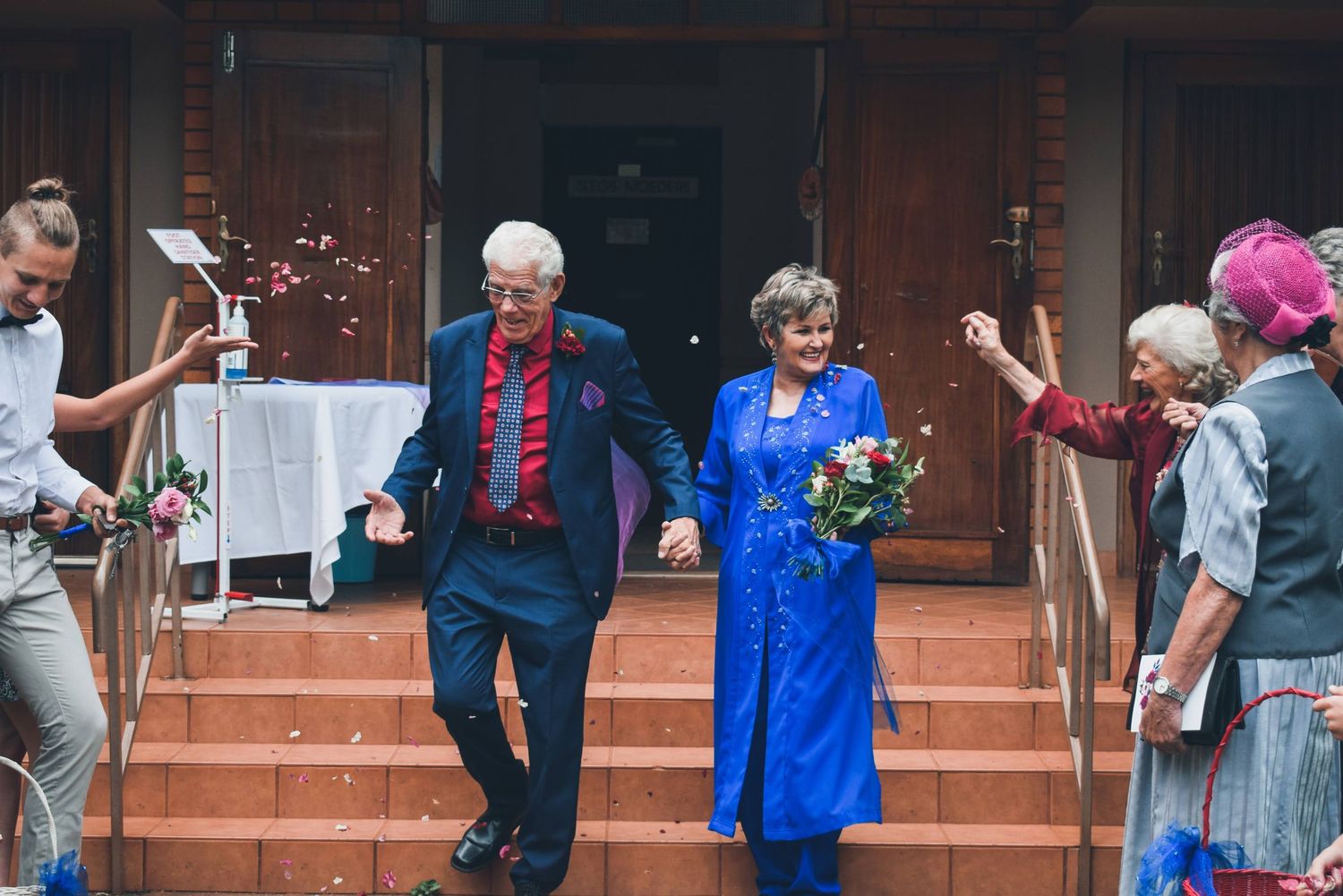 Elderly couple in blue and red outfits celebrating wedding exit with confetti and cheering guests on church steps.