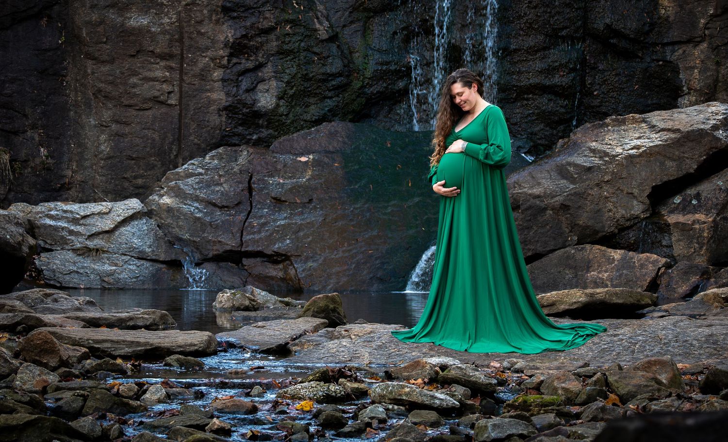 Pregnant woman in elegant green maternity gown standing on rocks near a waterfall in a serene outdoor setting.