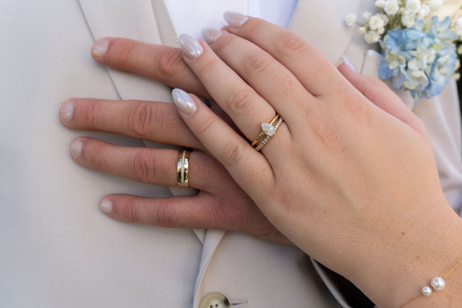 Newlywed couple displaying matching gold wedding rings on their hands, with a floral boutonniere in the background.