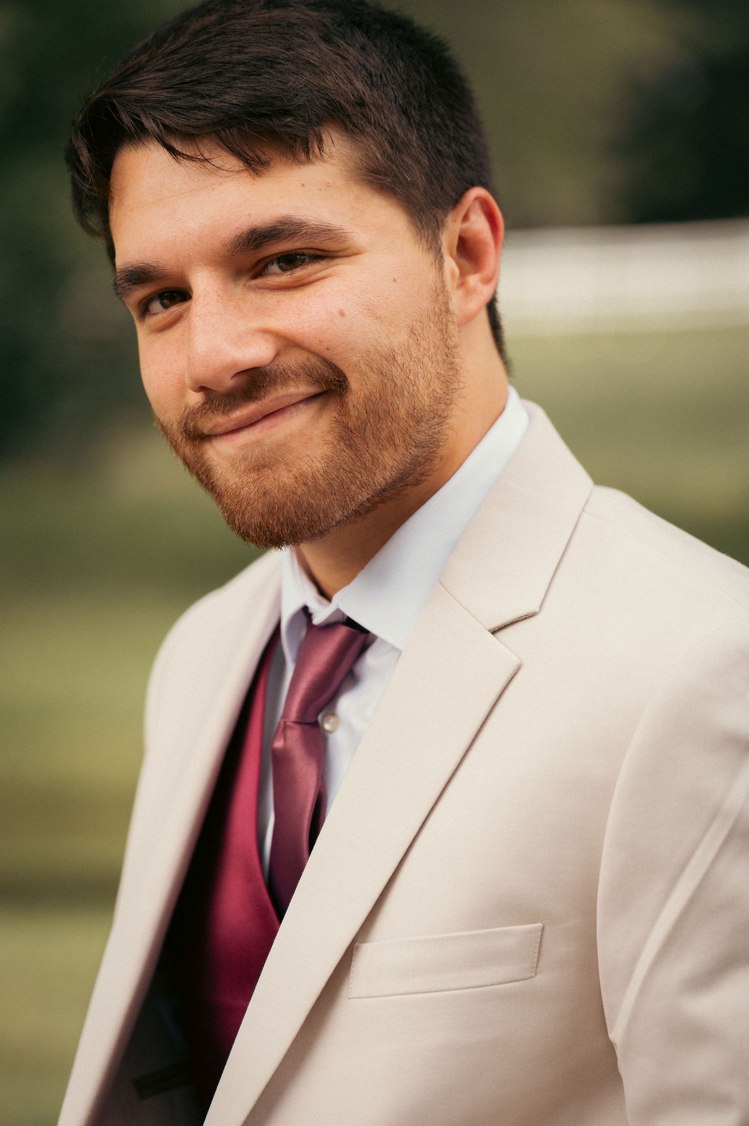 Handsome groom smiling in beige suit with burgundy tie outdoors.
