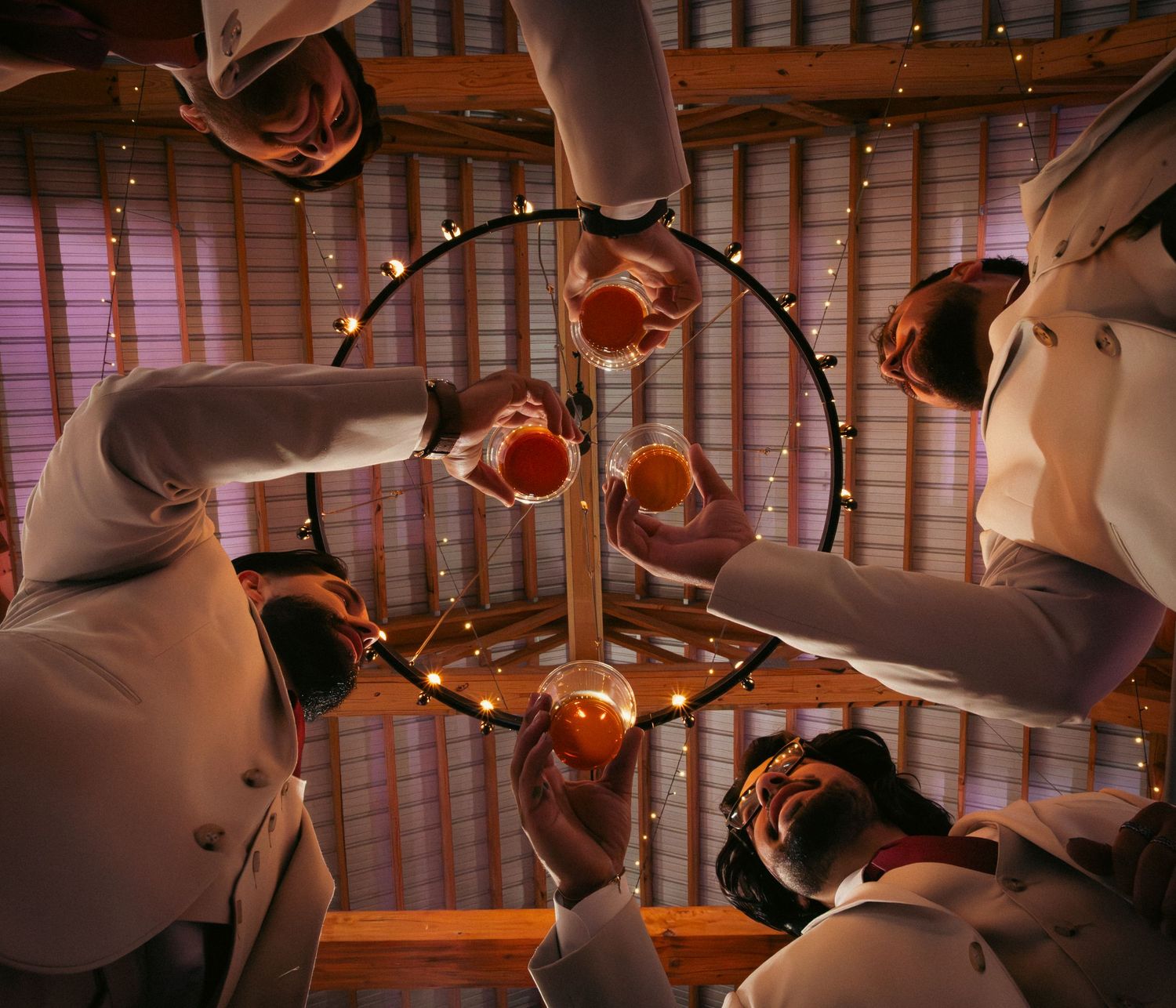 Wedding party toasting drinks beneath a chandelier, photographed from below in warm light.