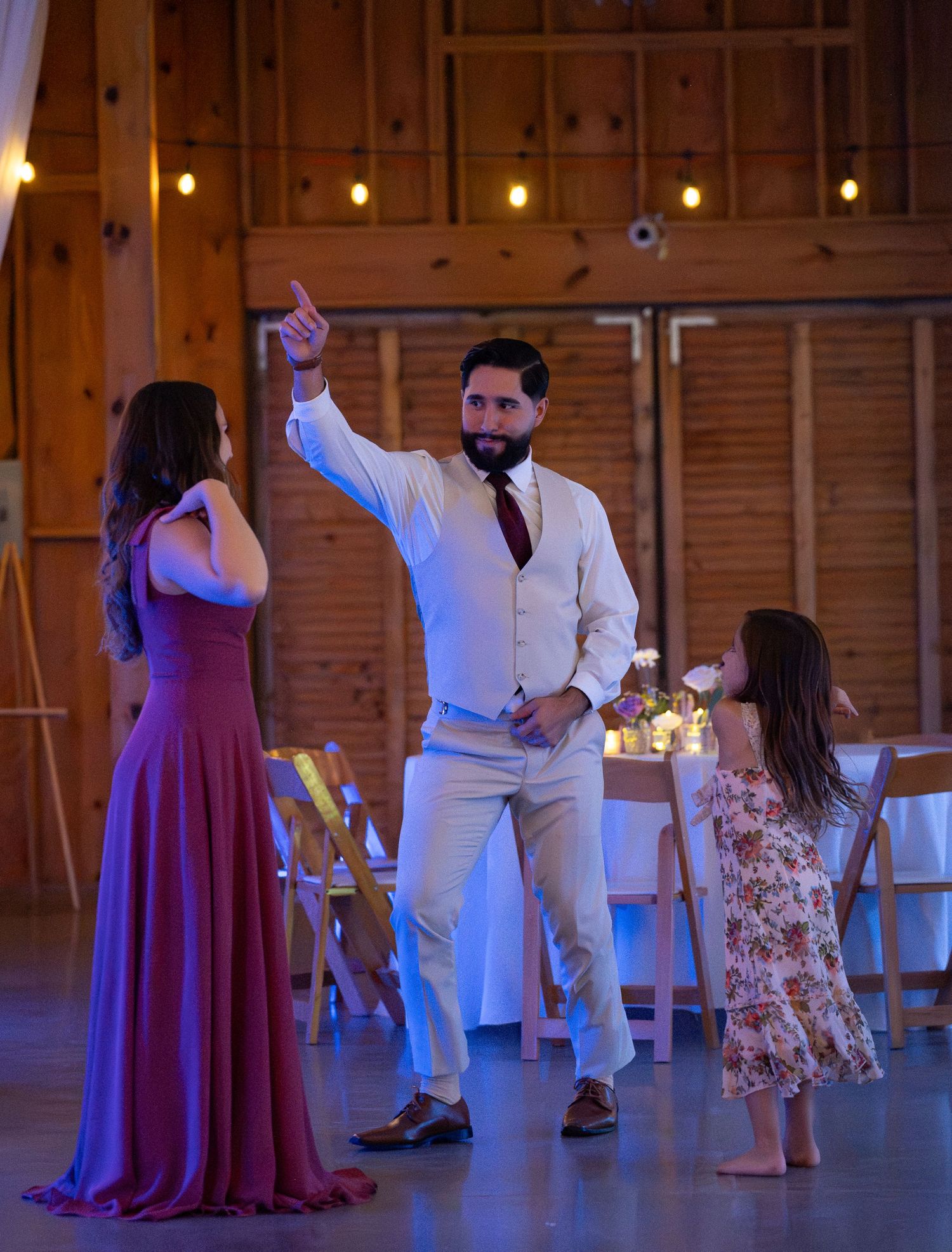 A groom in a white vest dances with a woman in a purple gown and a young girl in a floral dress at a barn wedding.